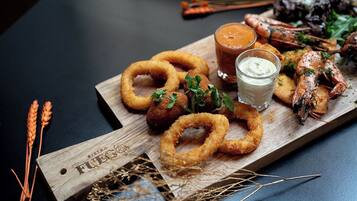 Ein Holzbrett mit gebratenen Speisen, darunter Zwiebelringe und Garnelen, sowie ein Glas Sauce.
