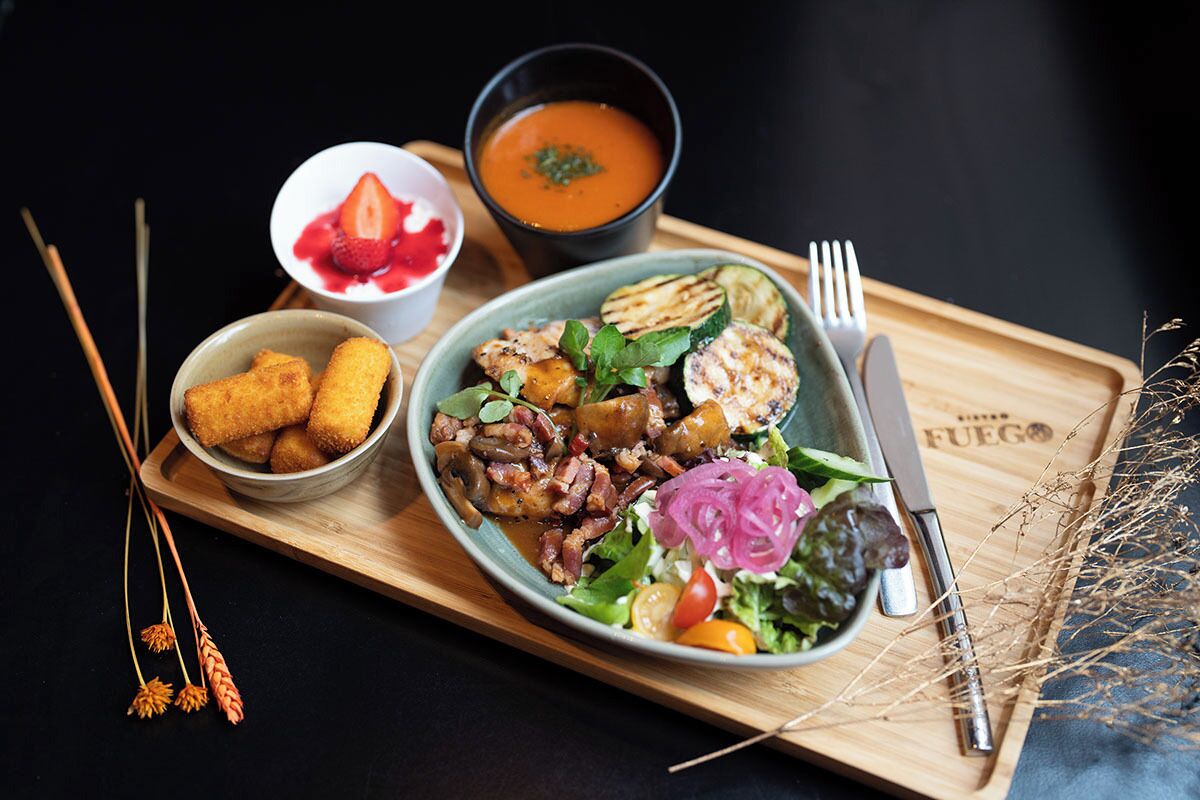 A meal with grilled chicken, vegetables, and a side of soup on a wooden board.