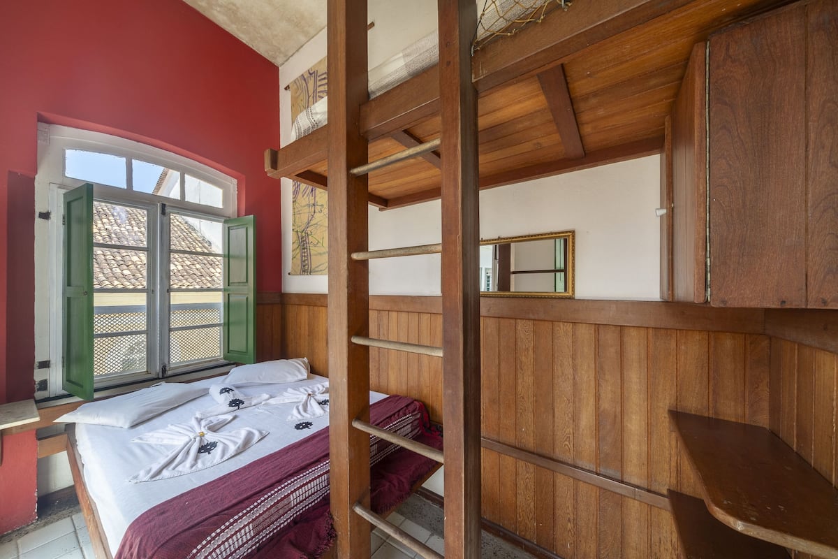 Ein Zimmer mit einem Bett, einem Fenster mit grünen Rollläden, einem Holzbettkasten und einem Spiegel an der Wand.