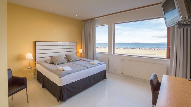 A hotel room with a bed, bedside tables, a TV, and a view of the beach.