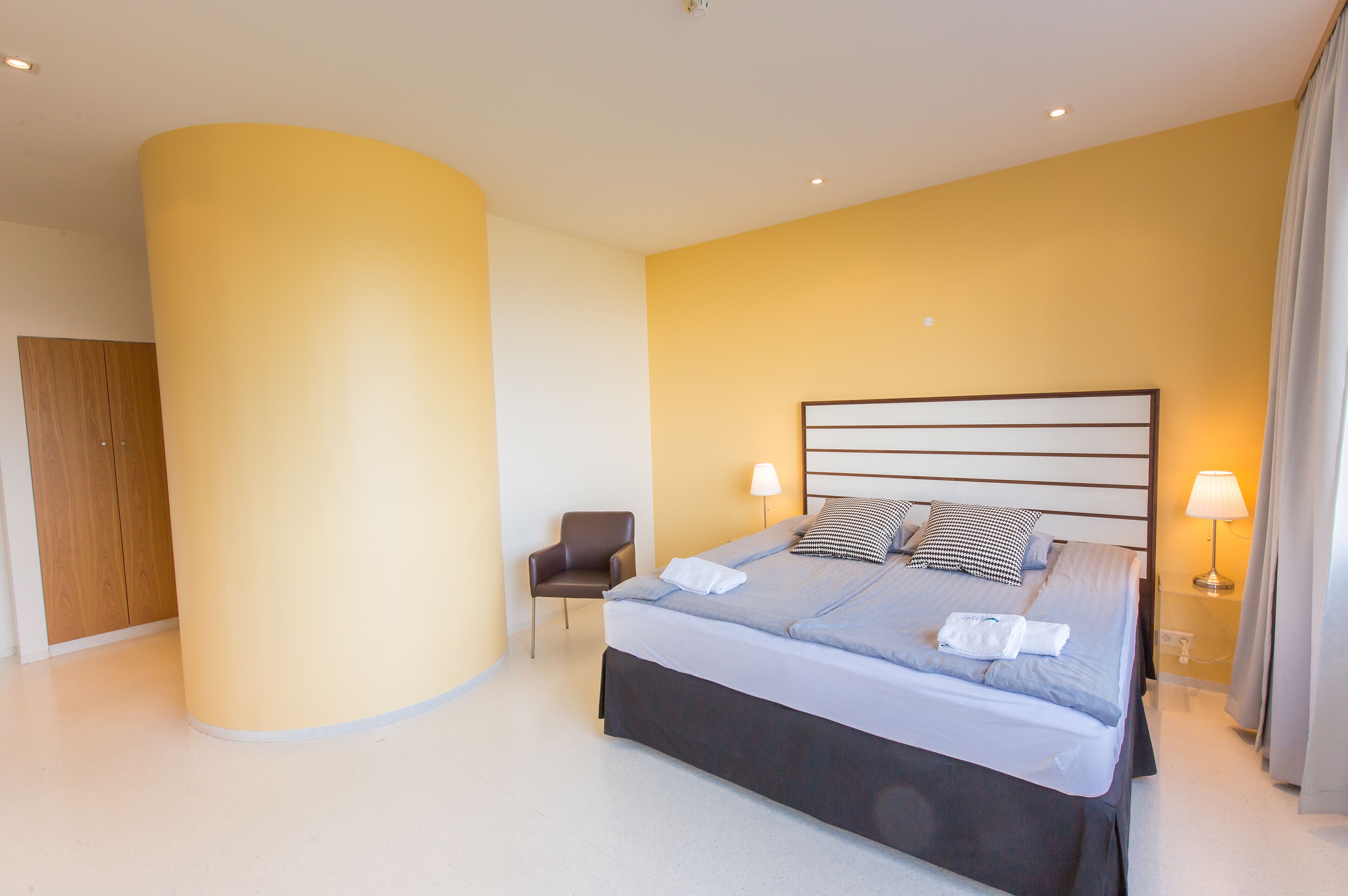 A modern hotel room with a large bed, a chair, and a wardrobe.