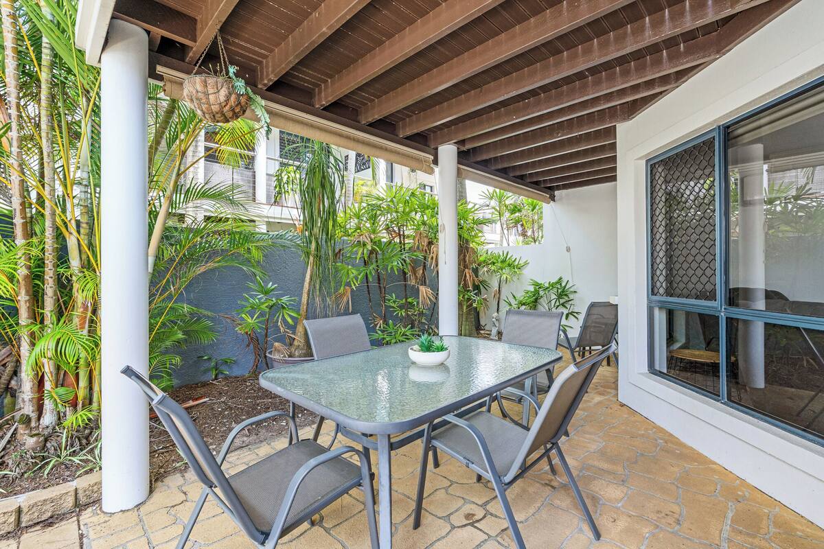 A covered patio with a table and chairs, surrounded by plants and a view of a building.