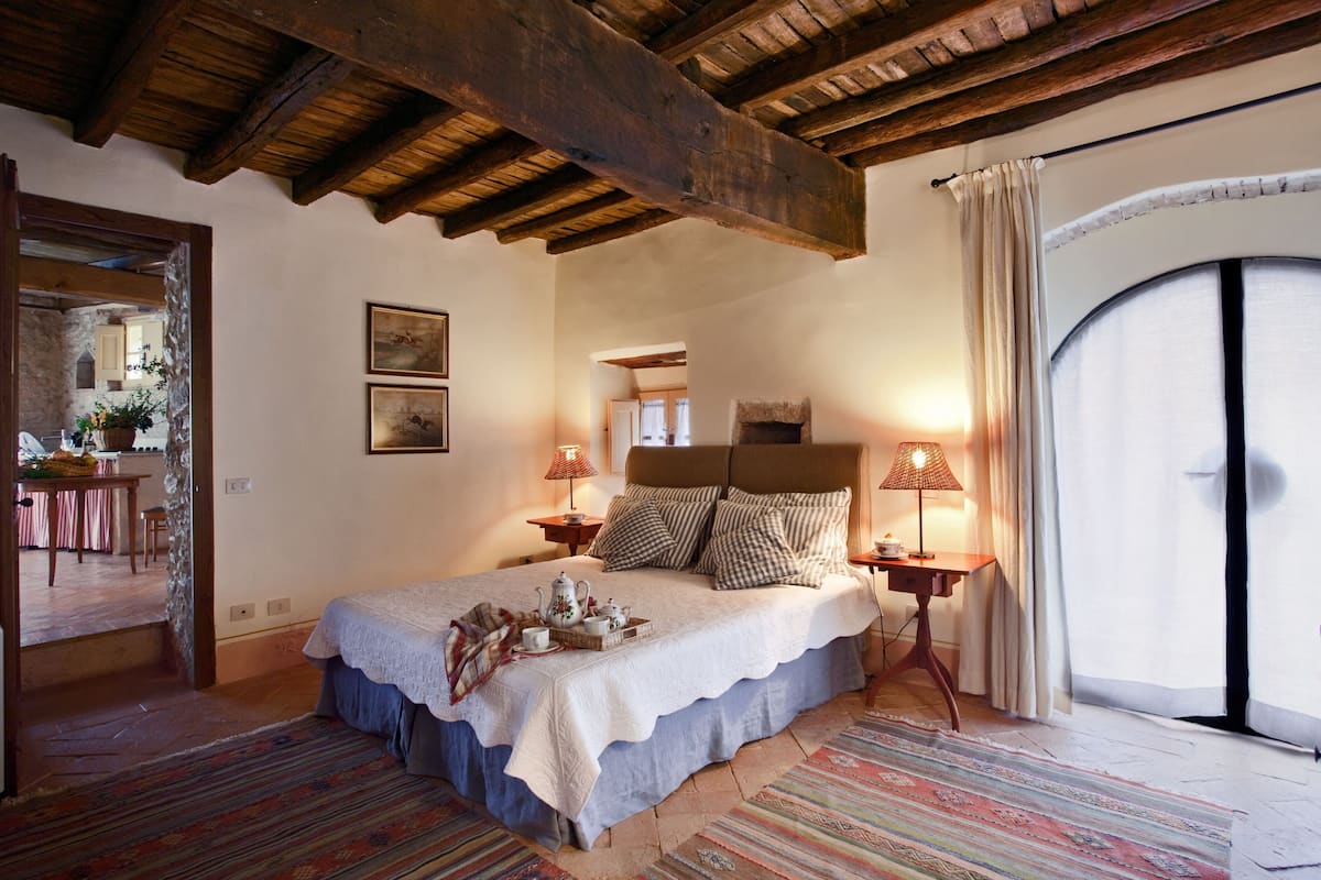 Ein gemütliches Schlafzimmer mit einem großen Bett, Holzbalken, einem kleinen Tisch mit Lampe und Blick ins Freie durch ein Fenster.
