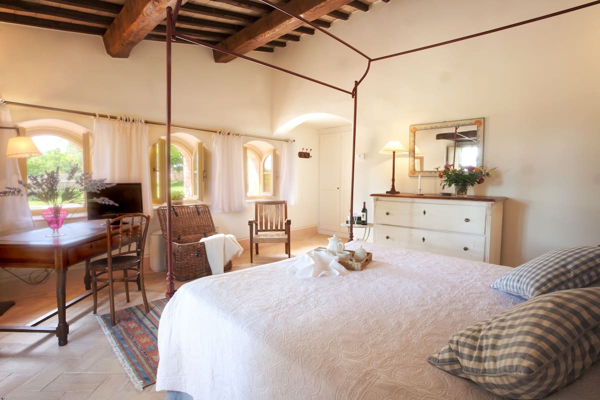 Ein Schlafzimmer mit einem großen Bett, einem Schminktisch, einem Sessel, einem Schreibtisch und einem Fenster mit Vorhängen.