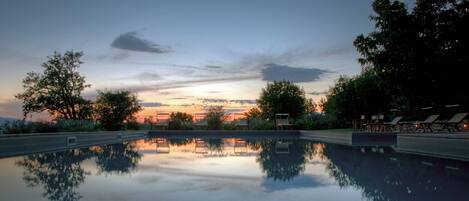Una piscina con un reflejo cristalino, rodeada de árboles y sillas de descanso, con un atardecer de fondo.