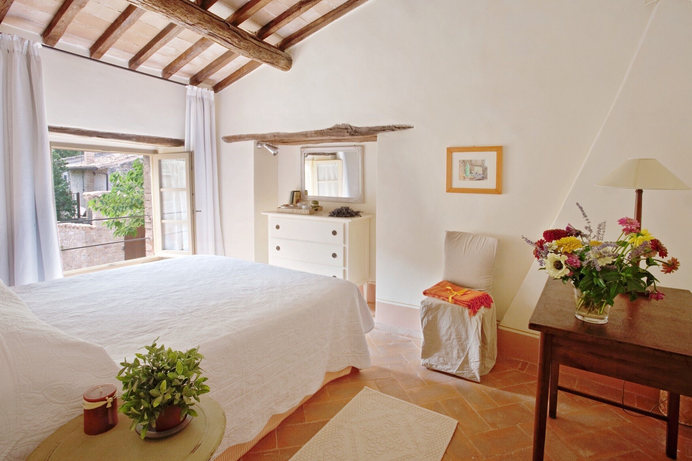 A bedroom with a bed, a dresser, a chair, a table with a vase of flowers, and a window with curtains.
