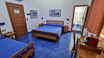 Habitación de hotel con dos camas, una puerta de madera, una mesita pequeña, una silla, un espejo y un baño visible a través de un vidrio.