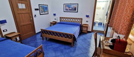 Habitación de hotel con dos camas, una puerta de madera, una mesita pequeña, una silla, un espejo y un baño visible a través de un vidrio.