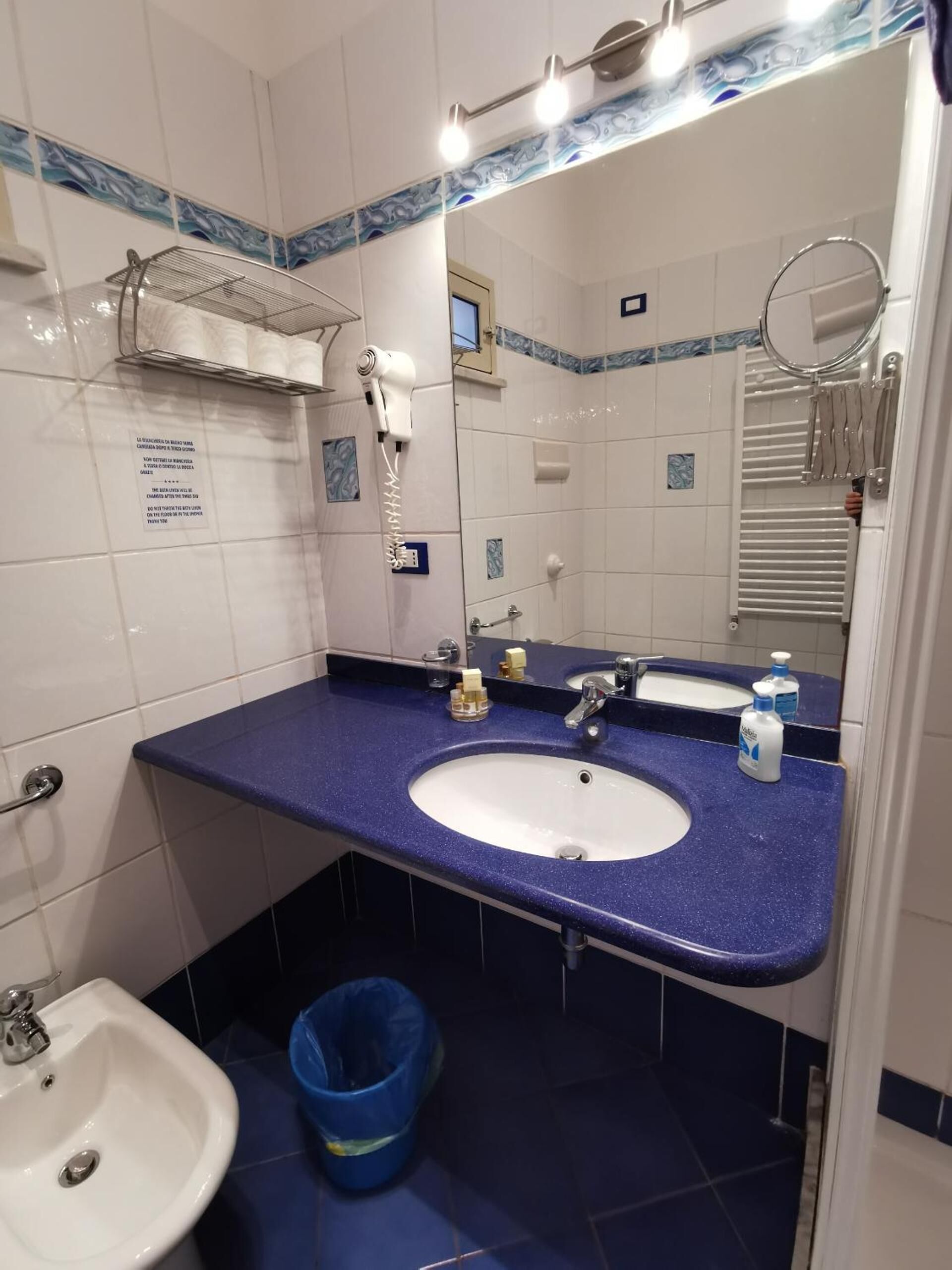 A bathroom with a white sink, blue countertop, and a mirror. There is a blue trash bin and a towel rack with towels.