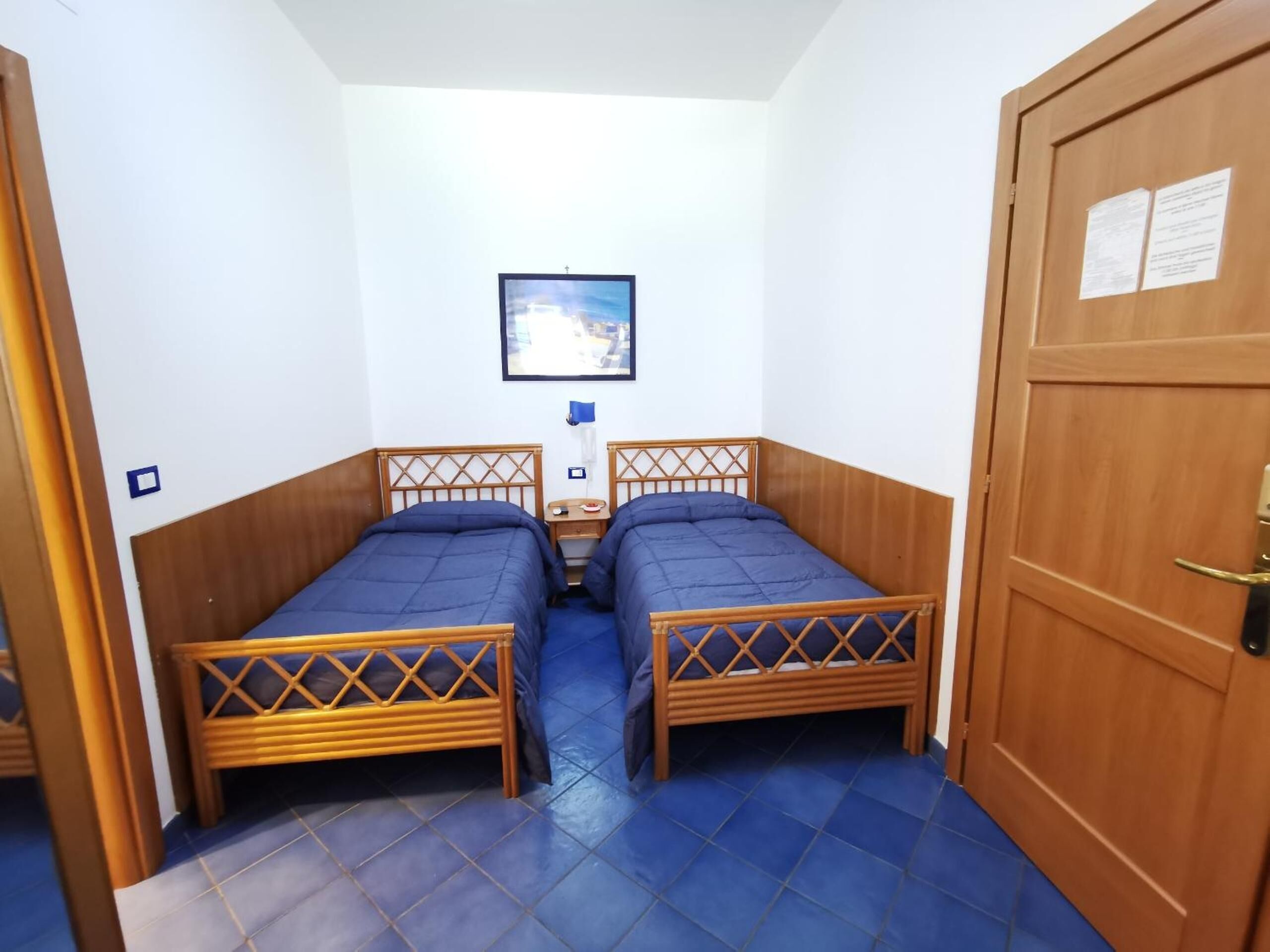 A small room with two wooden beds, a nightstand, a framed picture on the wall, and a door with a lock.