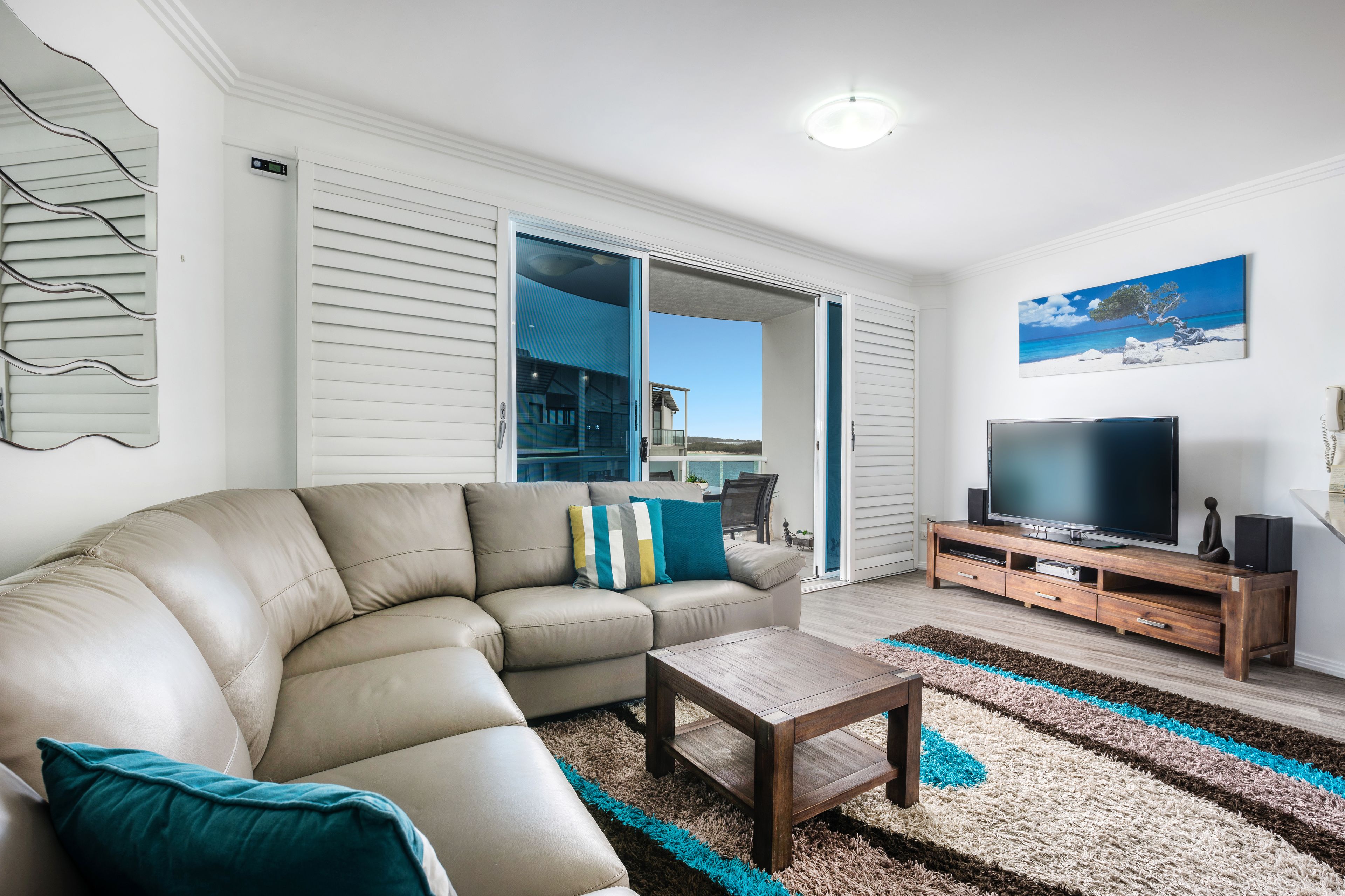 A living room with a beige sectional sofa, a wooden coffee table, a flat-screen TV on a wooden stand, and a large window offering a view of the outdoors.