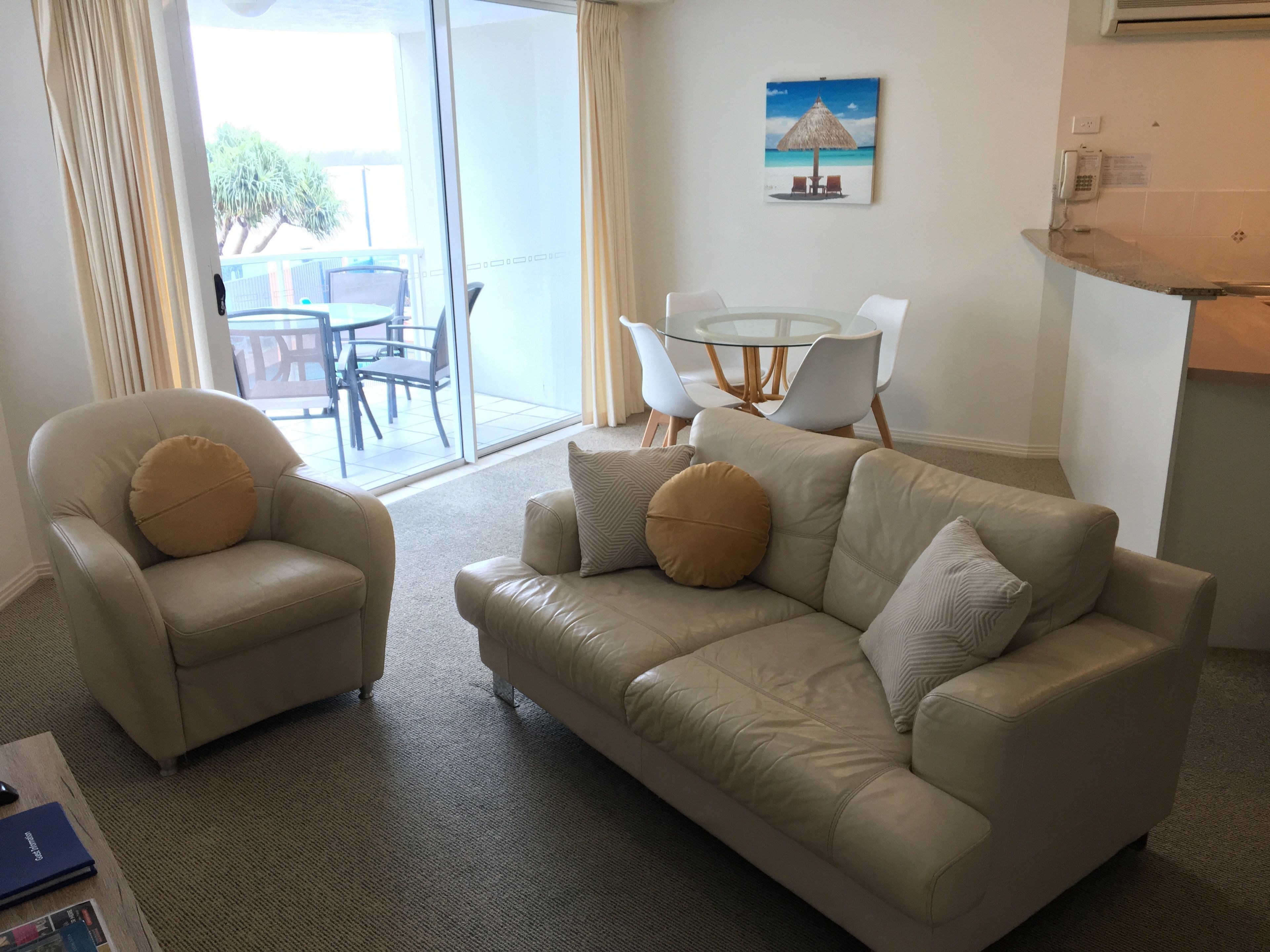 A living room with a sofa and armchair, a glass table with chairs, and a painting on the wall.