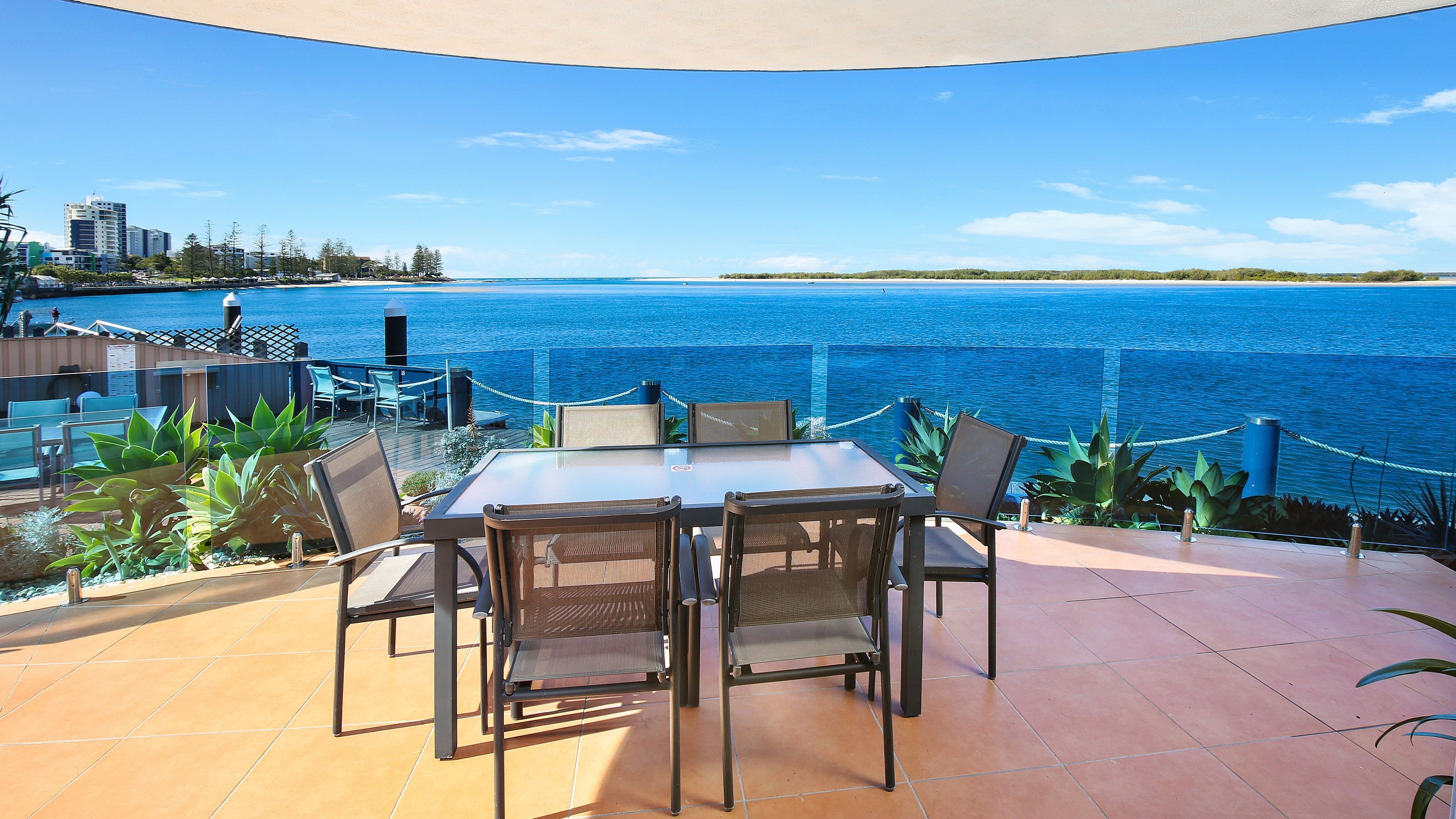 Una terraza con mesa y sillas que da a una masa de agua, con vista a la costa y edificios a lo lejos.