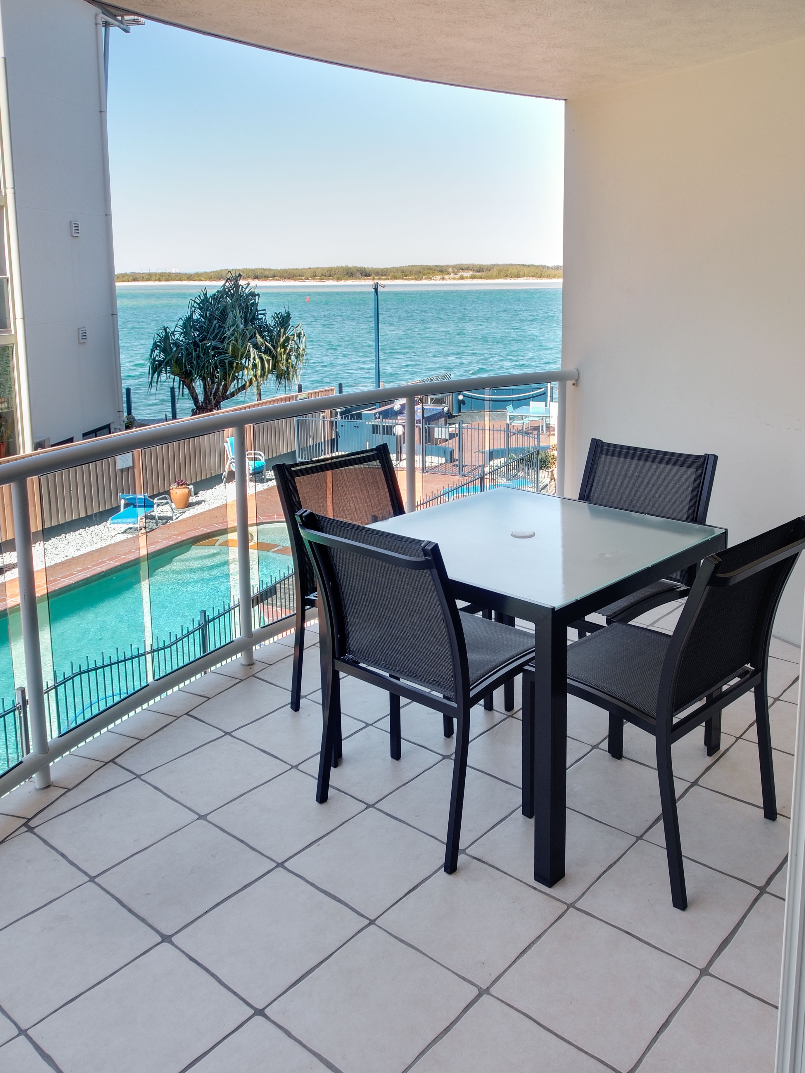 A balcony with a table and chairs, overlooking a pool and a beach.