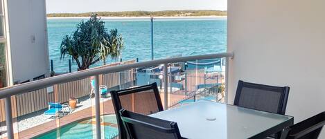 A balcony with a table and chairs, overlooking a pool and a beach.