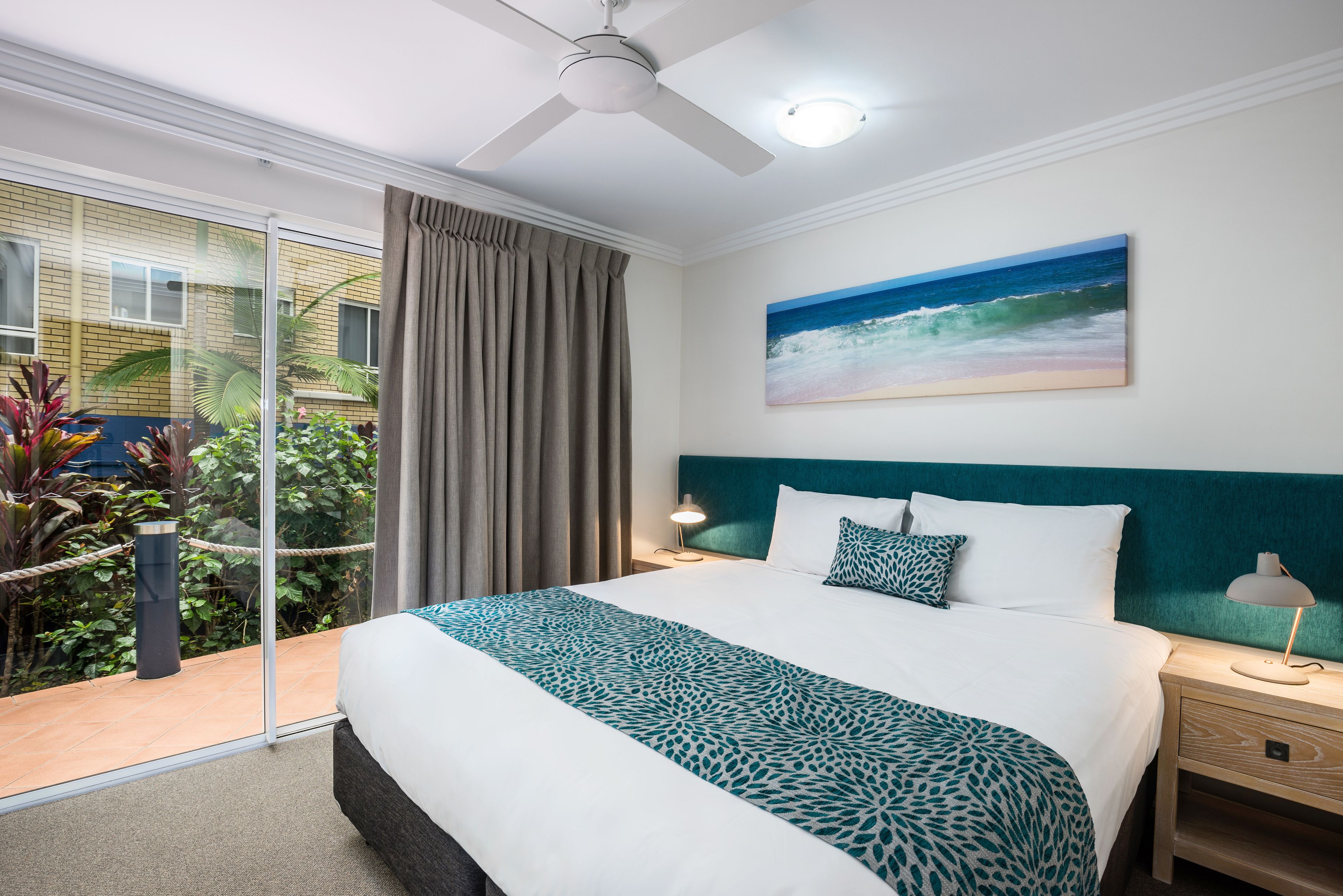 A hotel room with a bed, a ceiling fan, a nightstand, a lamp, a painting of the ocean, and a view of the outdoors.