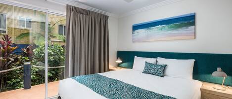 A hotel room with a bed, a ceiling fan, a nightstand, a lamp, a painting of the ocean, and a view of the outdoors.