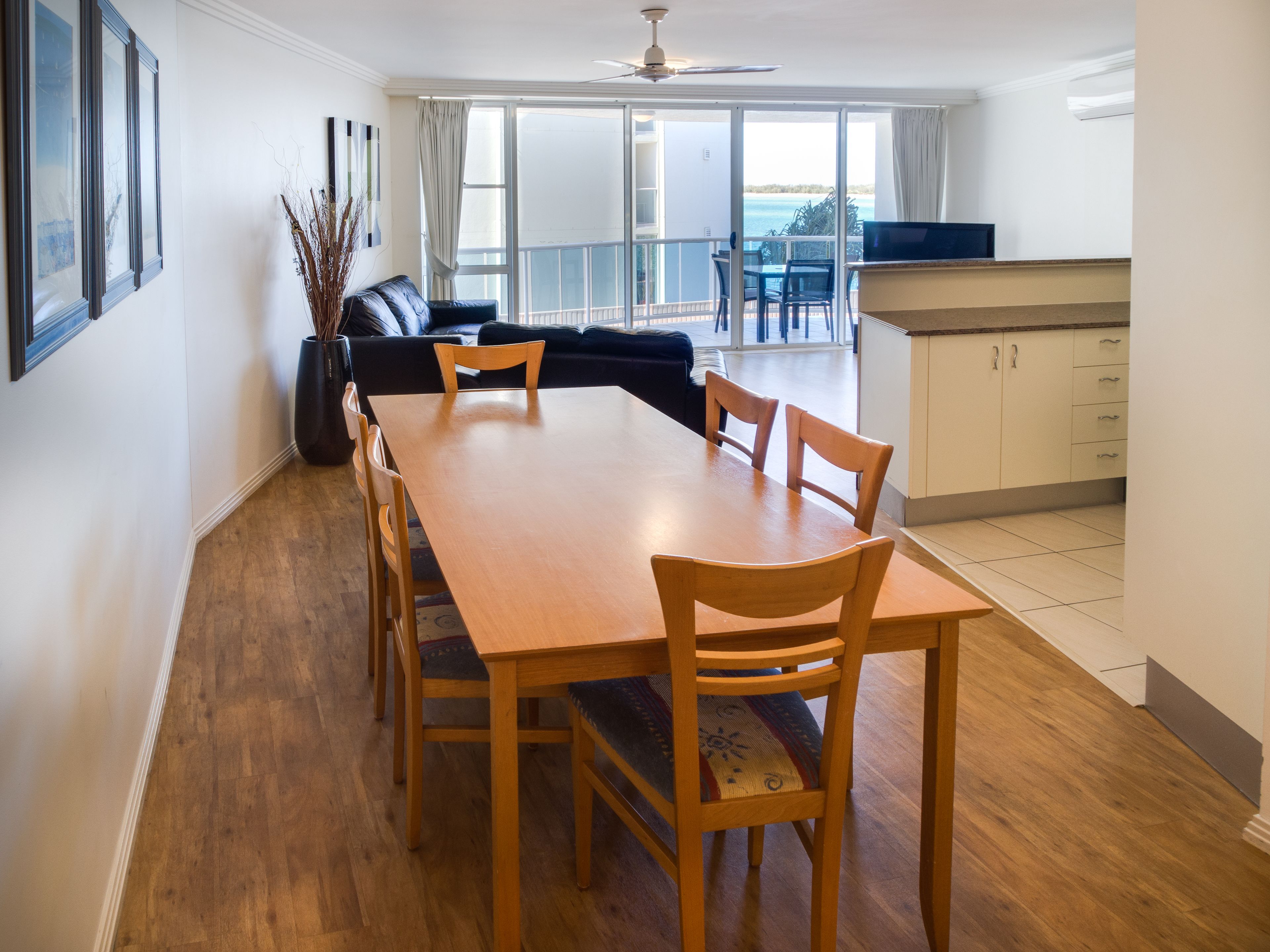 A spacious dining area with a long wooden table and six chairs, a black sofa, and a view of the ocean through large sliding doors.
