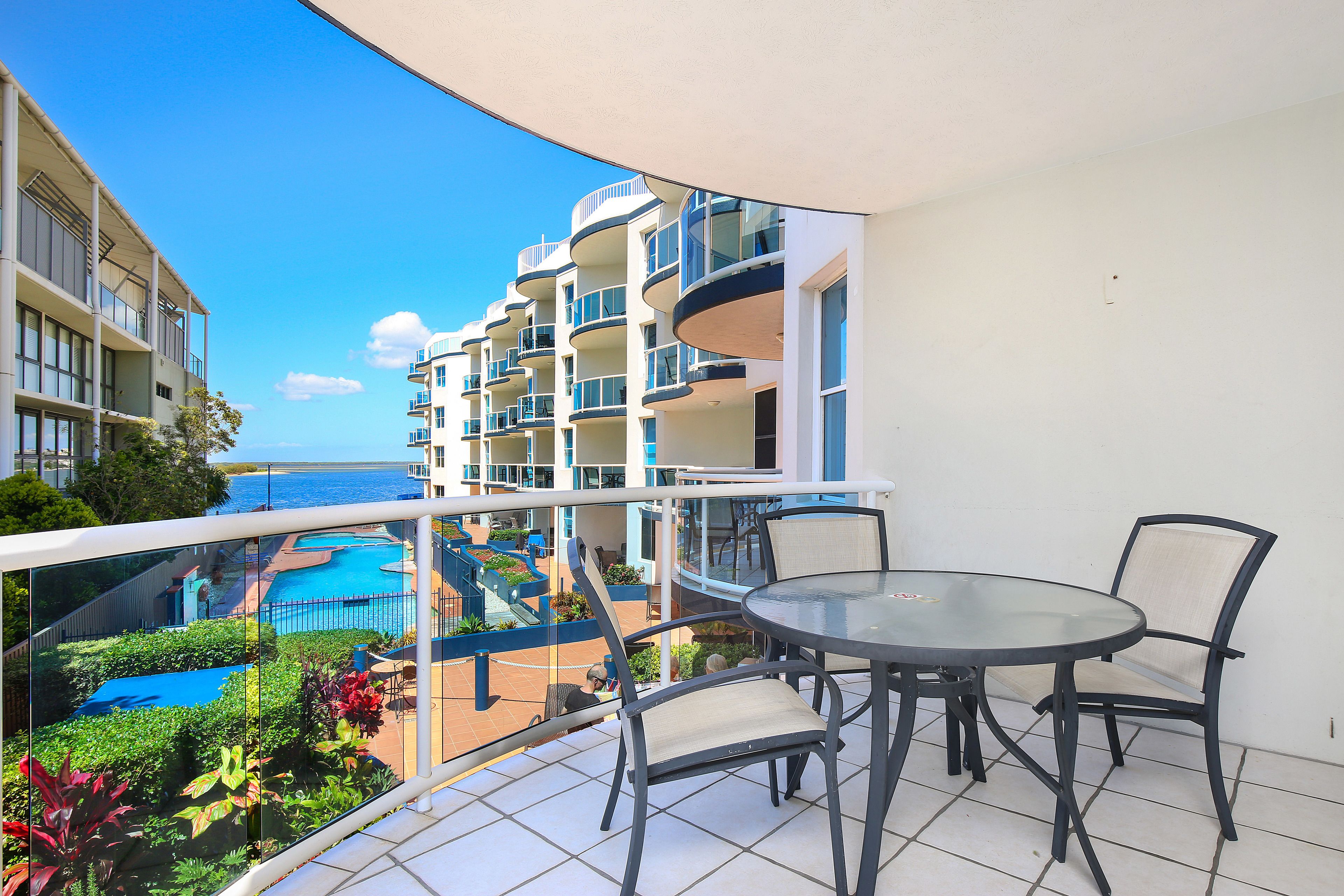 A balcony with a glass table and chairs, overlooking a pool and a building with multiple balconies.