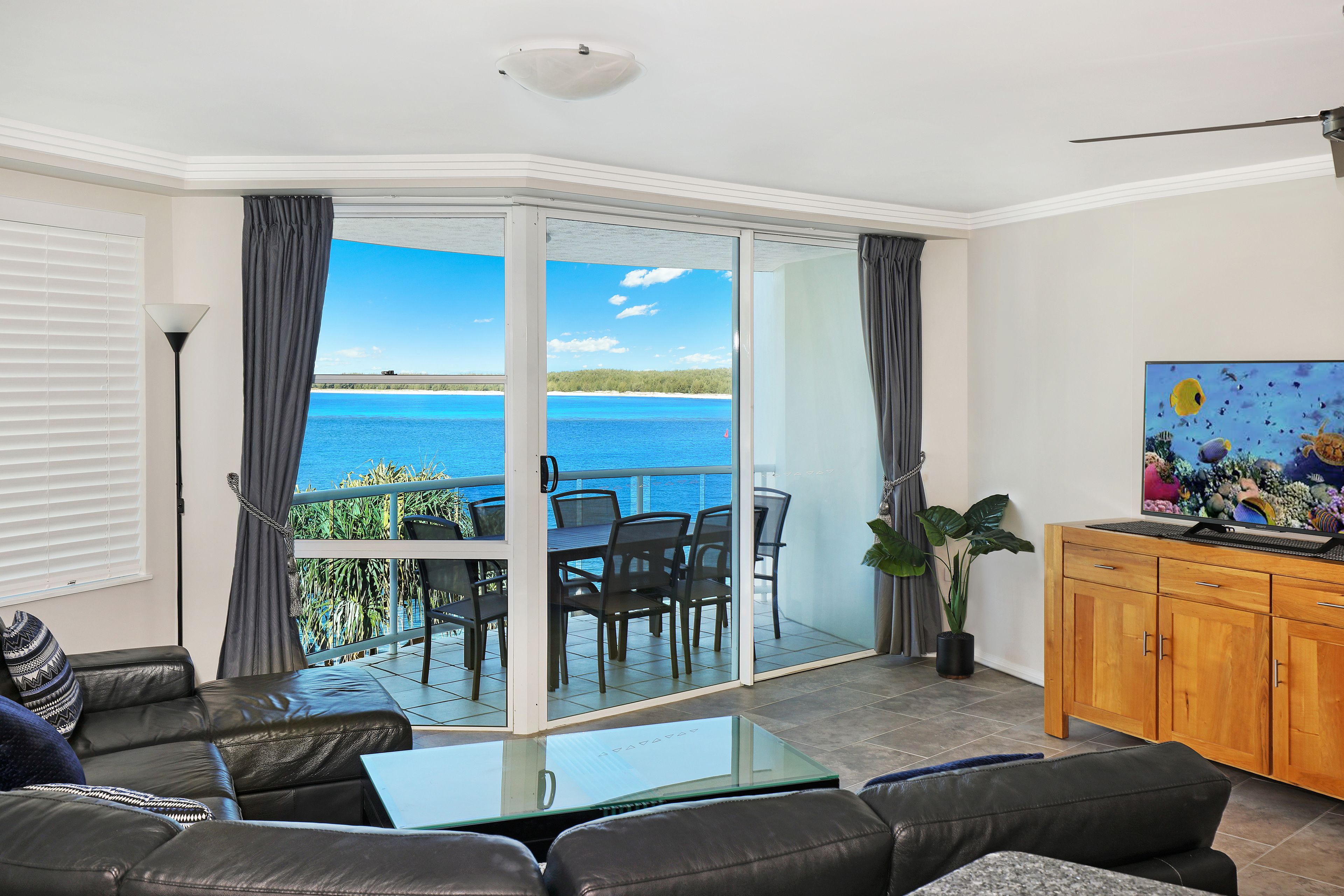 Habitación con balcón con vistas a un cuerpo de agua, un área de comedor y un televisor sobre un mueble de madera.