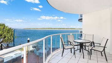 A balcony with a glass railing overlooking a body of water, a table with chairs, and a view of the coastline.