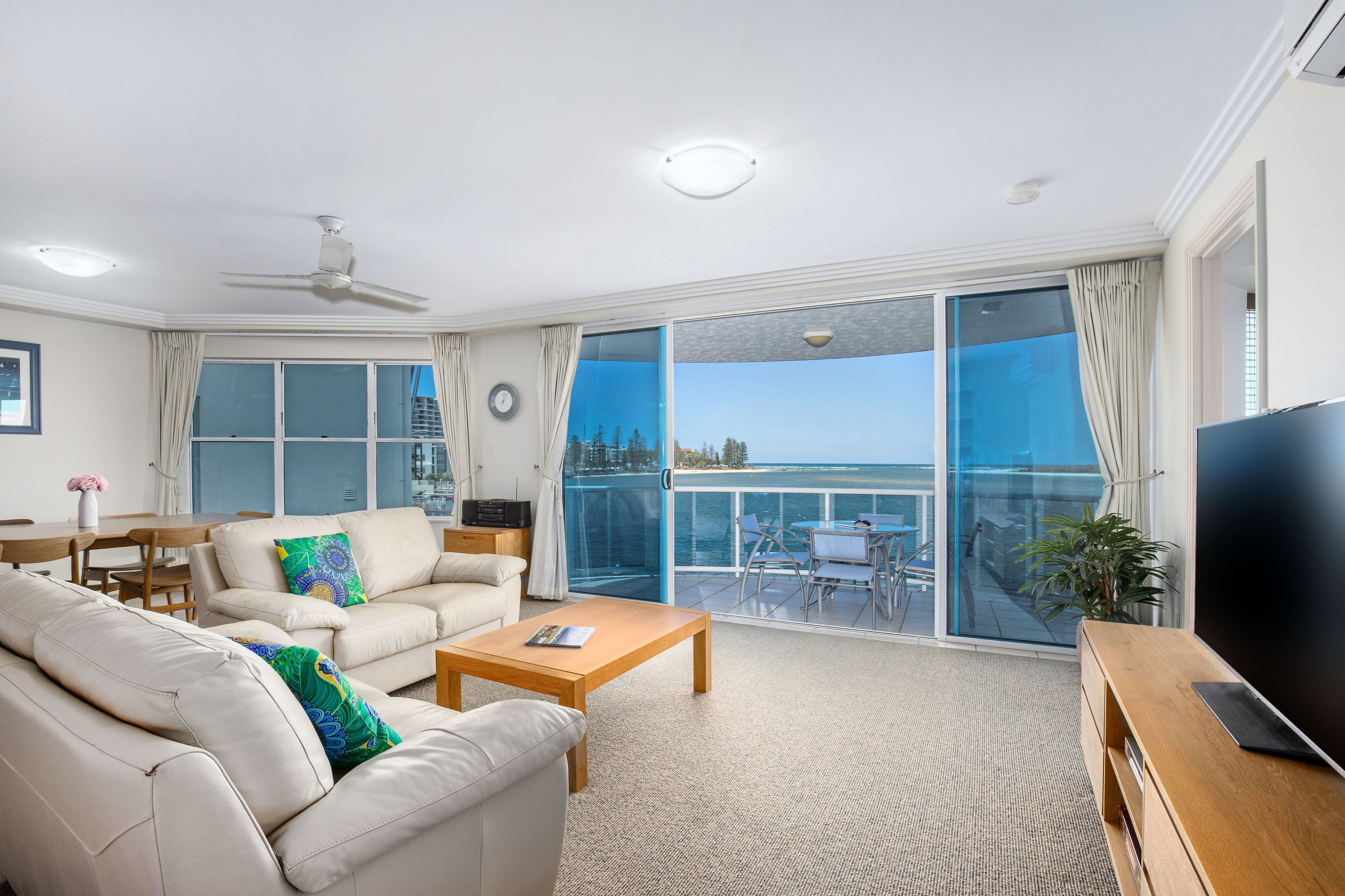 Un salón moderno con sofá, mesa de centro y televisor. Hay un balcón con vistas al mar.