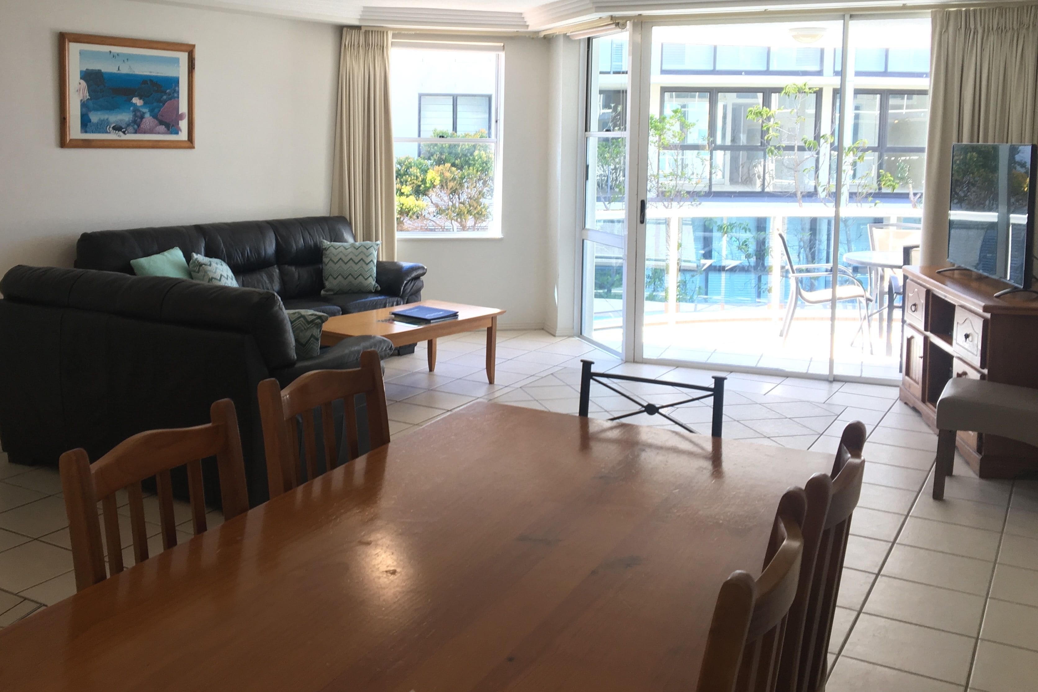 A living room with a sofa, coffee table, and dining table. There is a balcony with a pool and outdoor furniture.