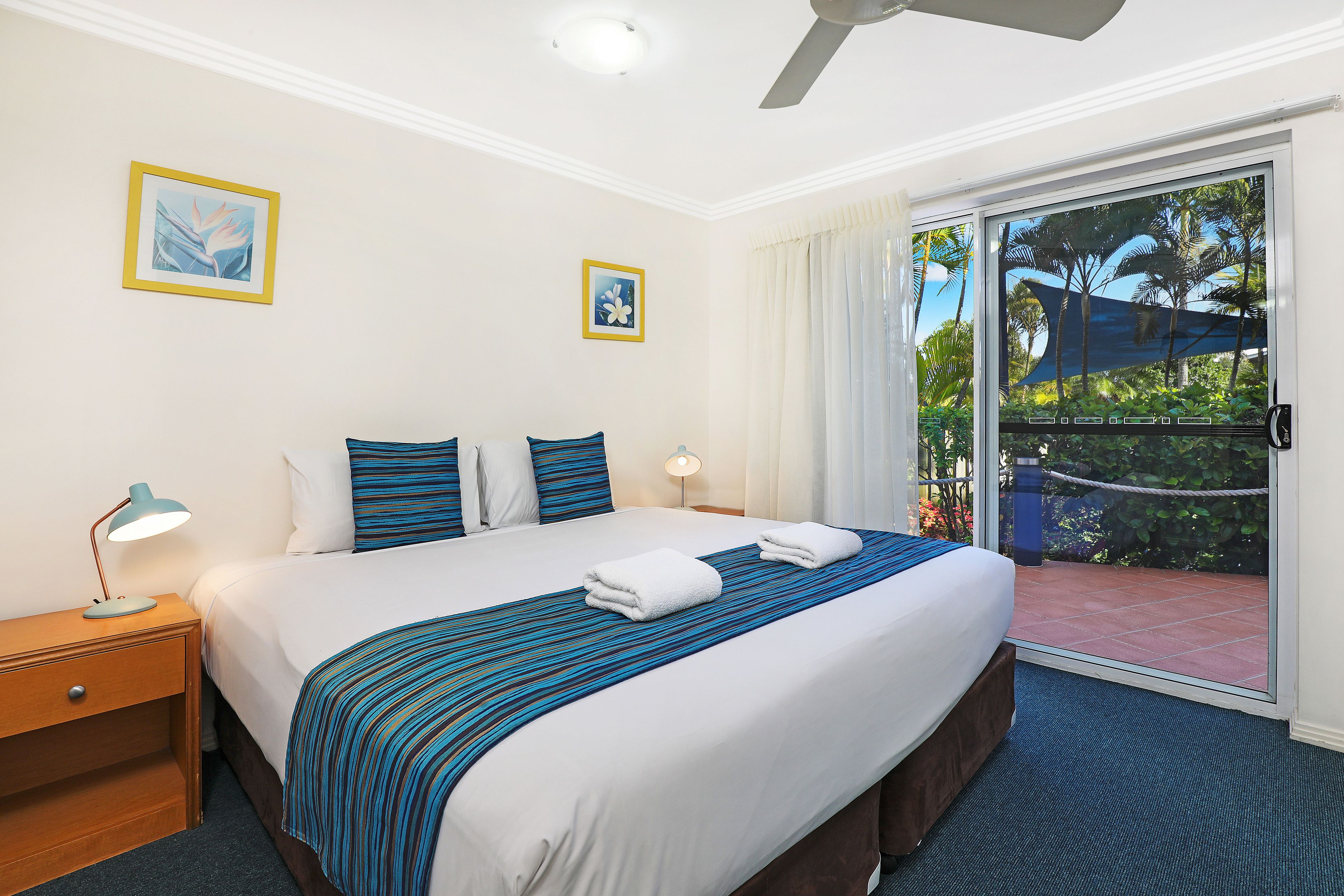 A hotel room with a large bed, a nightstand, a desk lamp, and a view of the outdoor area through a sliding glass door.