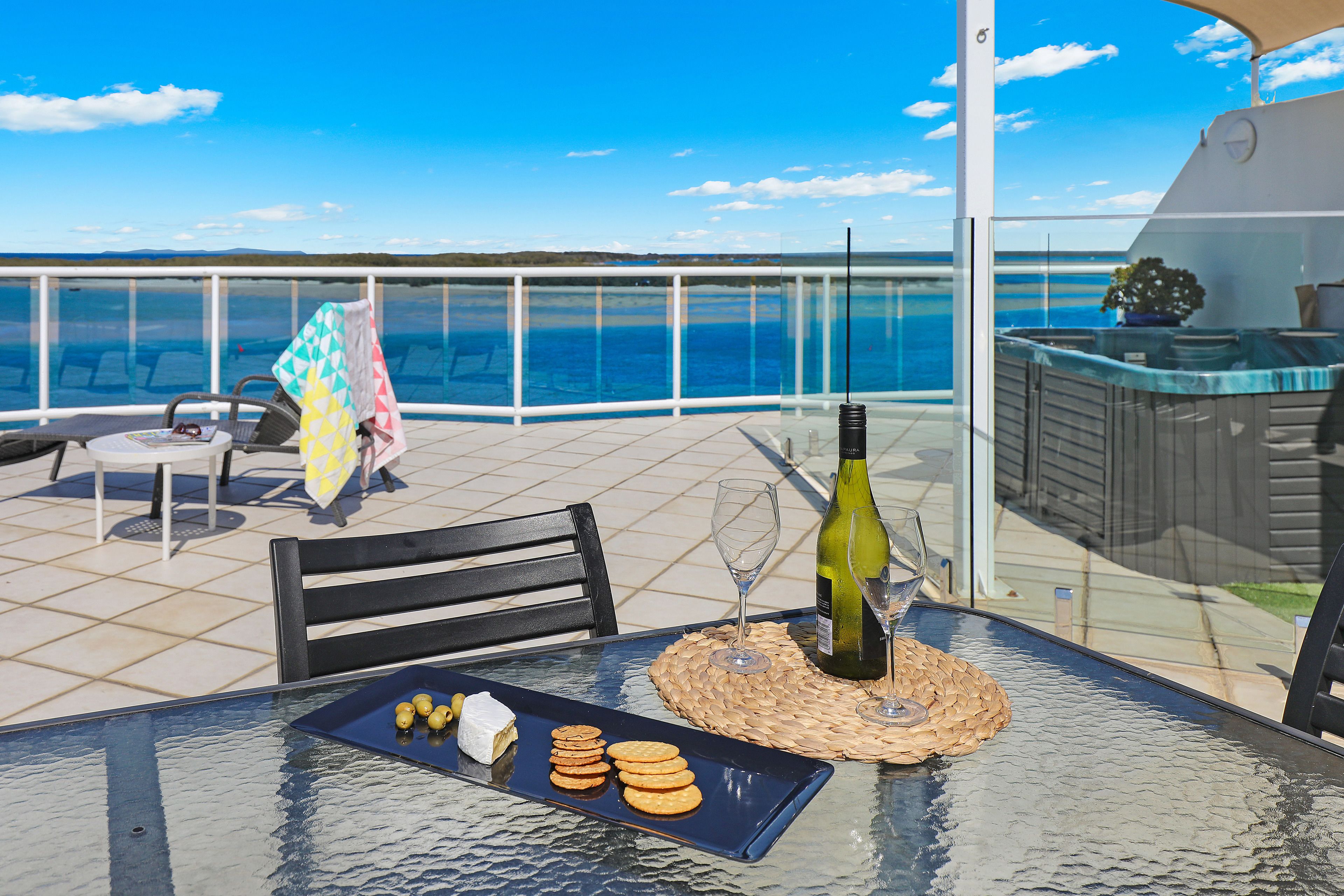 A glass-top table with a bottle of wine, glasses, and snacks on a balcony with a view of the sea.