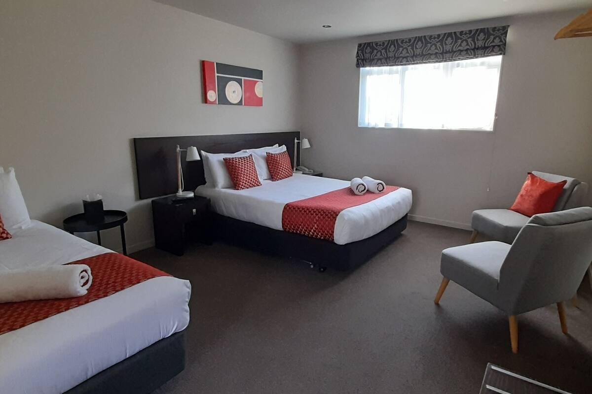 A hotel room with two beds, a chair, a nightstand, a lamp, and a window with curtains.