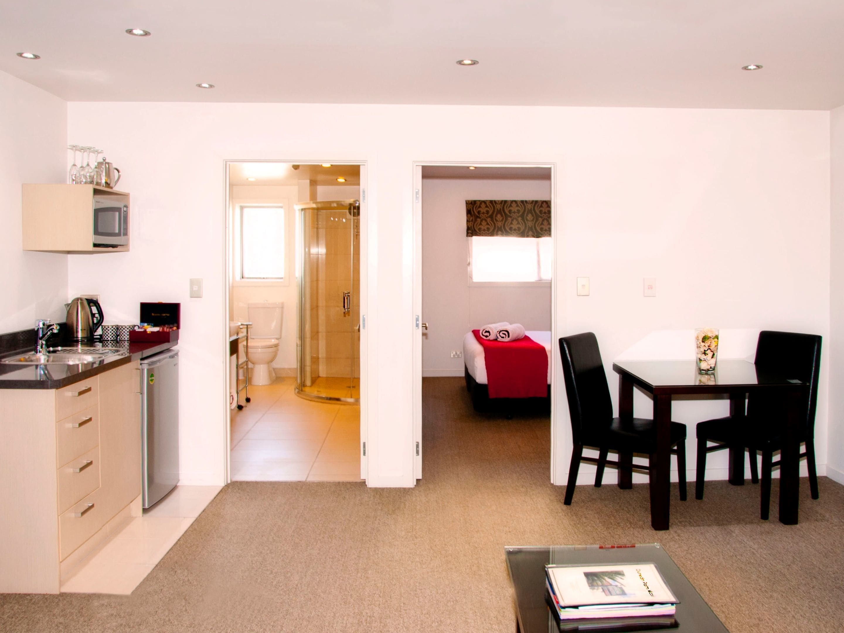 Une chambre d’hôtel compacte comprenant un coin cuisine, une douche, un lit avec une couverture rouge, un coin repas avec une table et des chaises, et un petit coin salon avec une table basse.