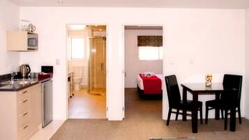 Une chambre d’hôtel compacte comprenant un coin cuisine, une douche, un lit avec une couverture rouge, un coin repas avec une table et des chaises, et un petit coin salon avec une table basse.