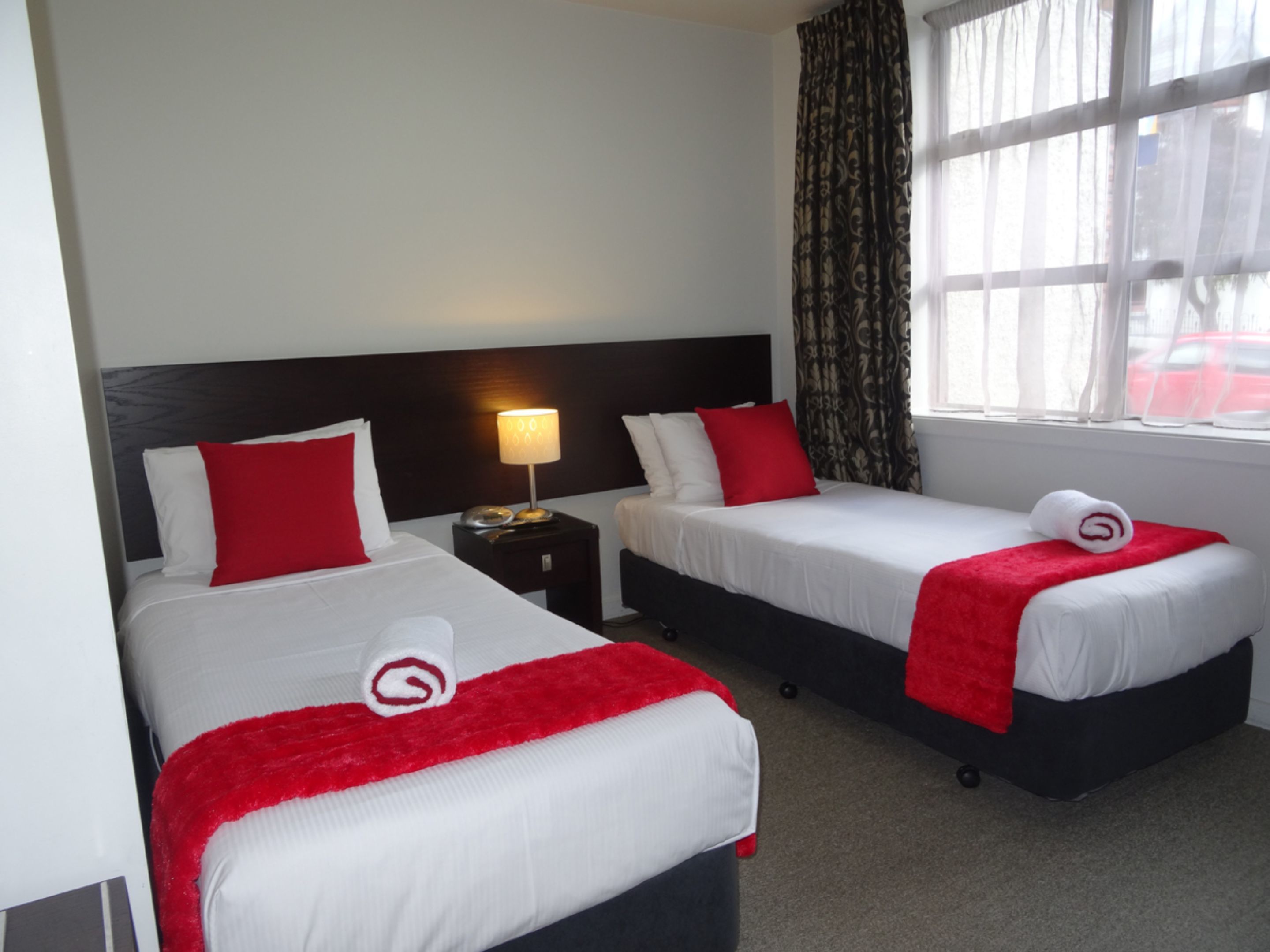 A hotel room with two beds, each with white bedding and red accents, a nightstand with a lamp, and a window with patterned curtains.