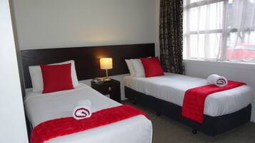 A hotel room with two beds, each with white bedding and red accents, a nightstand with a lamp, and a window with patterned curtains.