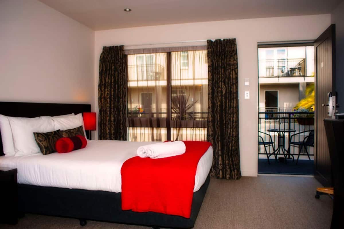 A hotel room with a bed, a nightstand, a red throw, and a balcony with outdoor seating.
