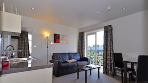 A modern living room with a sofa, coffee table, and dining area. There is a kitchen with a sink and cabinets. The room has large windows with curtains. - Dunedin Palms Motel (Dunedin)