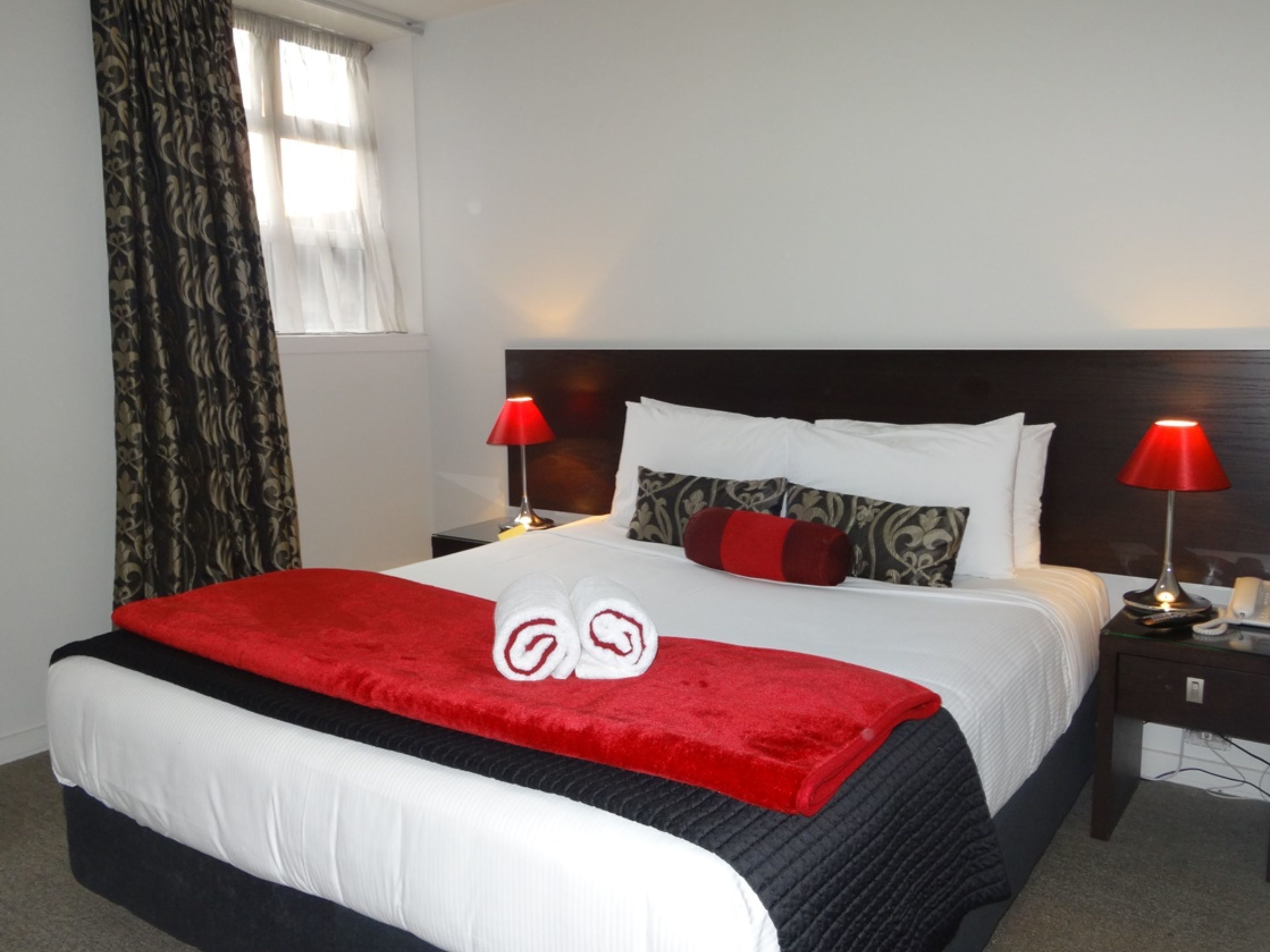 A neatly made bed with a red blanket and two towels folded on top. There are two bedside lamps with red shades, a bedside table with a phone and a remote control, and a window with curtains.
