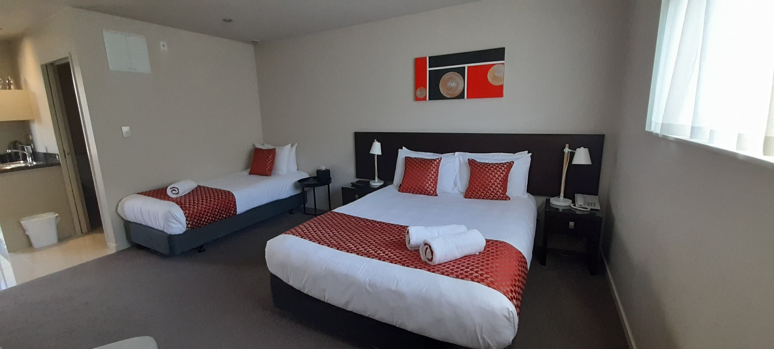 A hotel room with two beds, each with white linens and red accents, a small bedside table with a lamp, and a wall-mounted artwork above the beds.