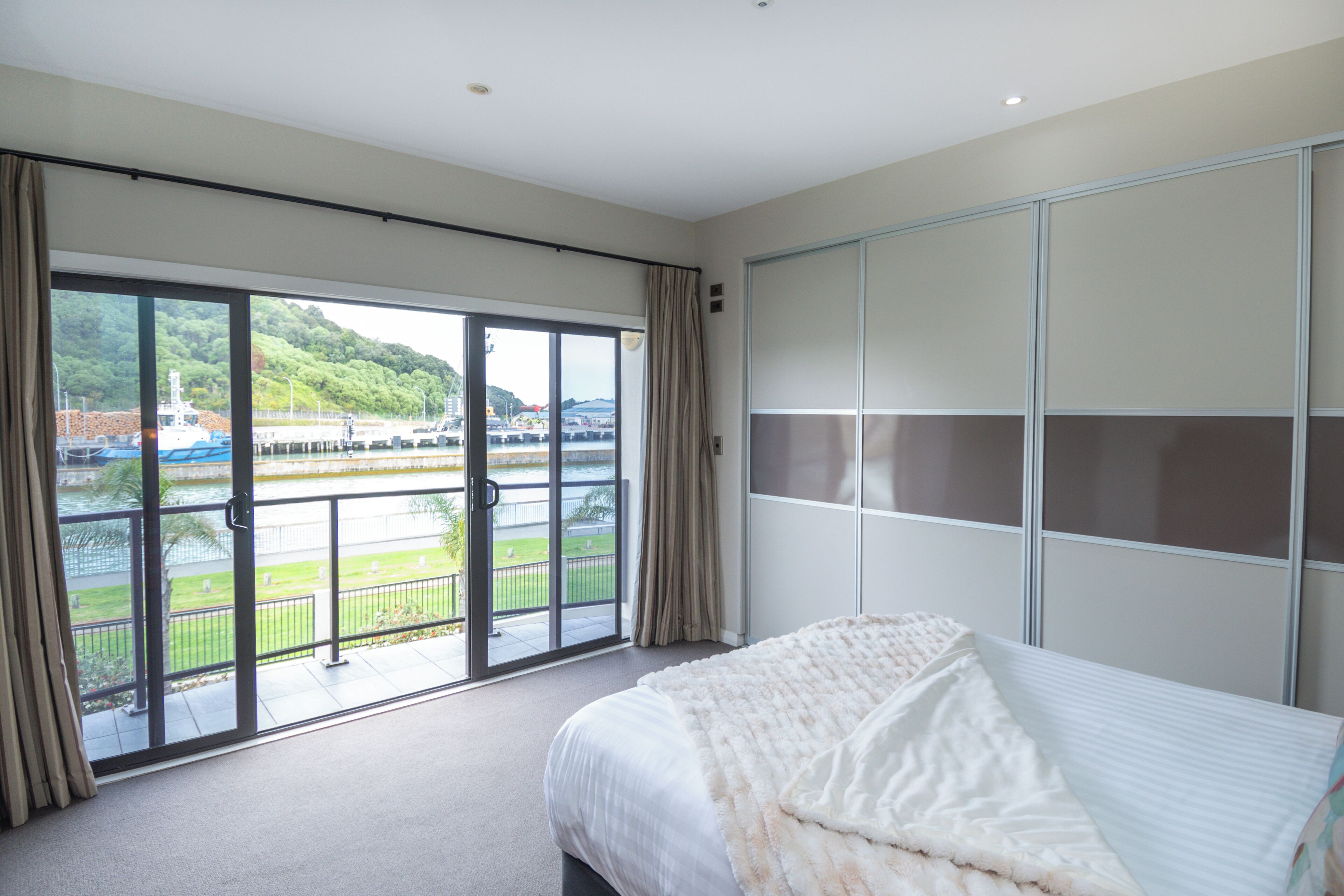 Quarto de hotel com uma grande porta de vidro deslizante que dá para uma varanda com vista para a água e vegetação.