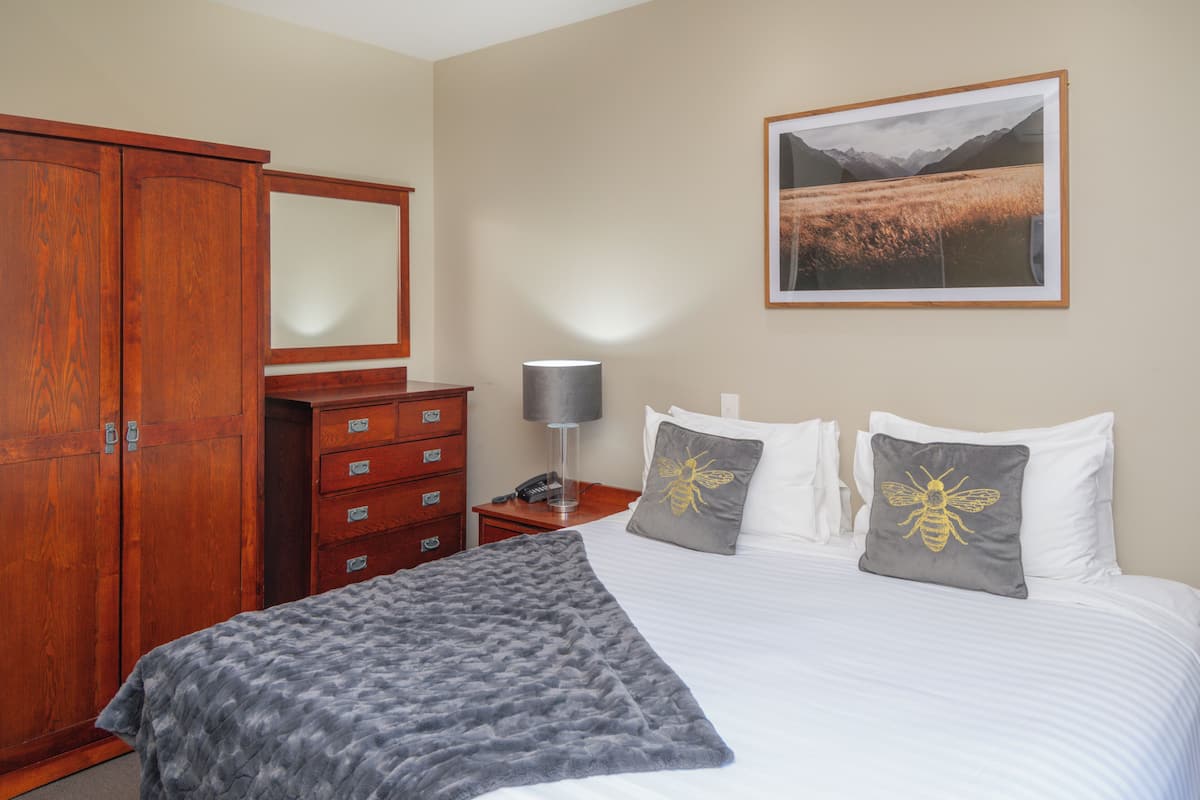 A hotel room with a bed, a wooden wardrobe, a desk with a lamp, and a framed landscape picture on the wall.