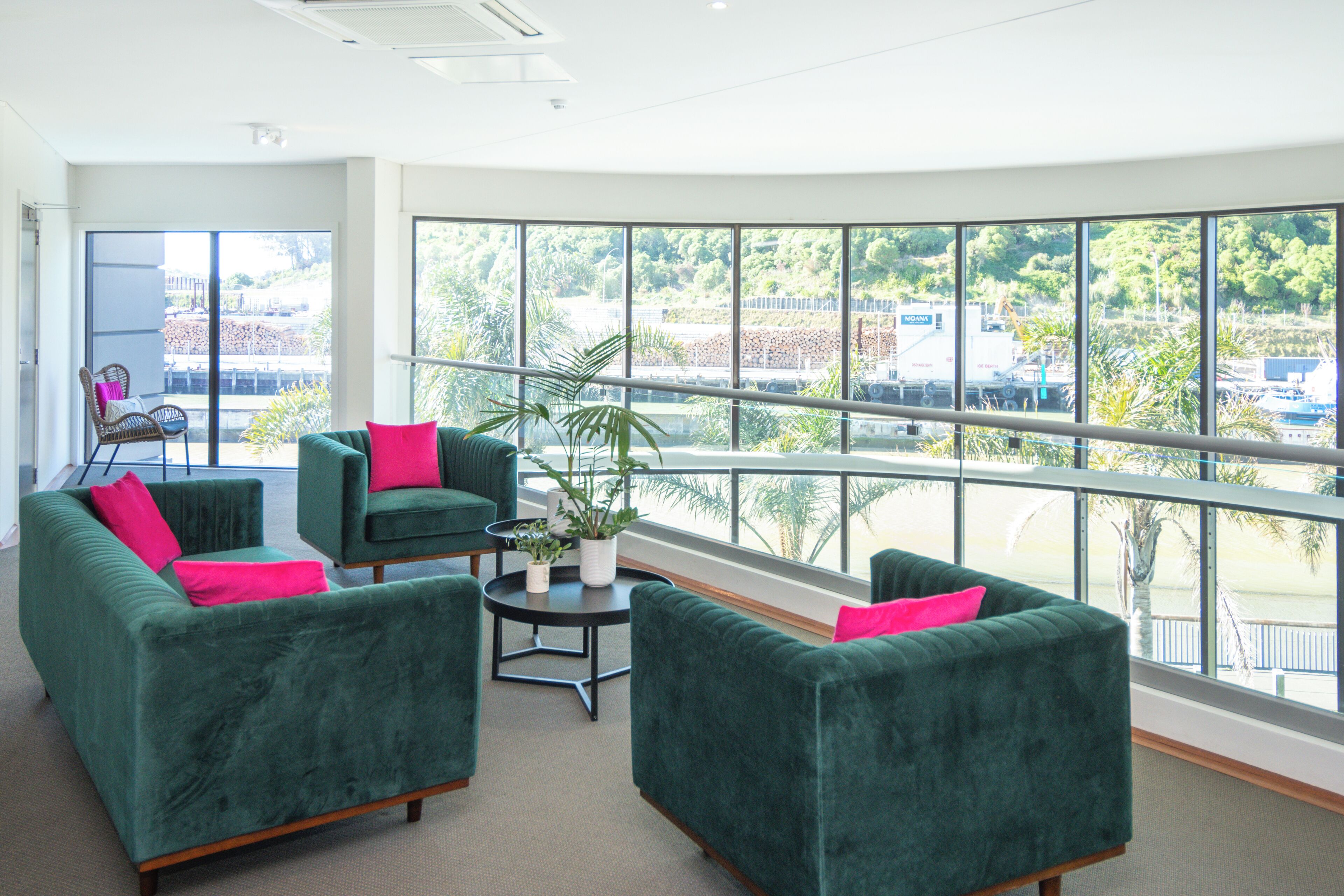 A spacious room with large windows, green sofas with pink cushions, a small table with a plant, and a view of a parking area.