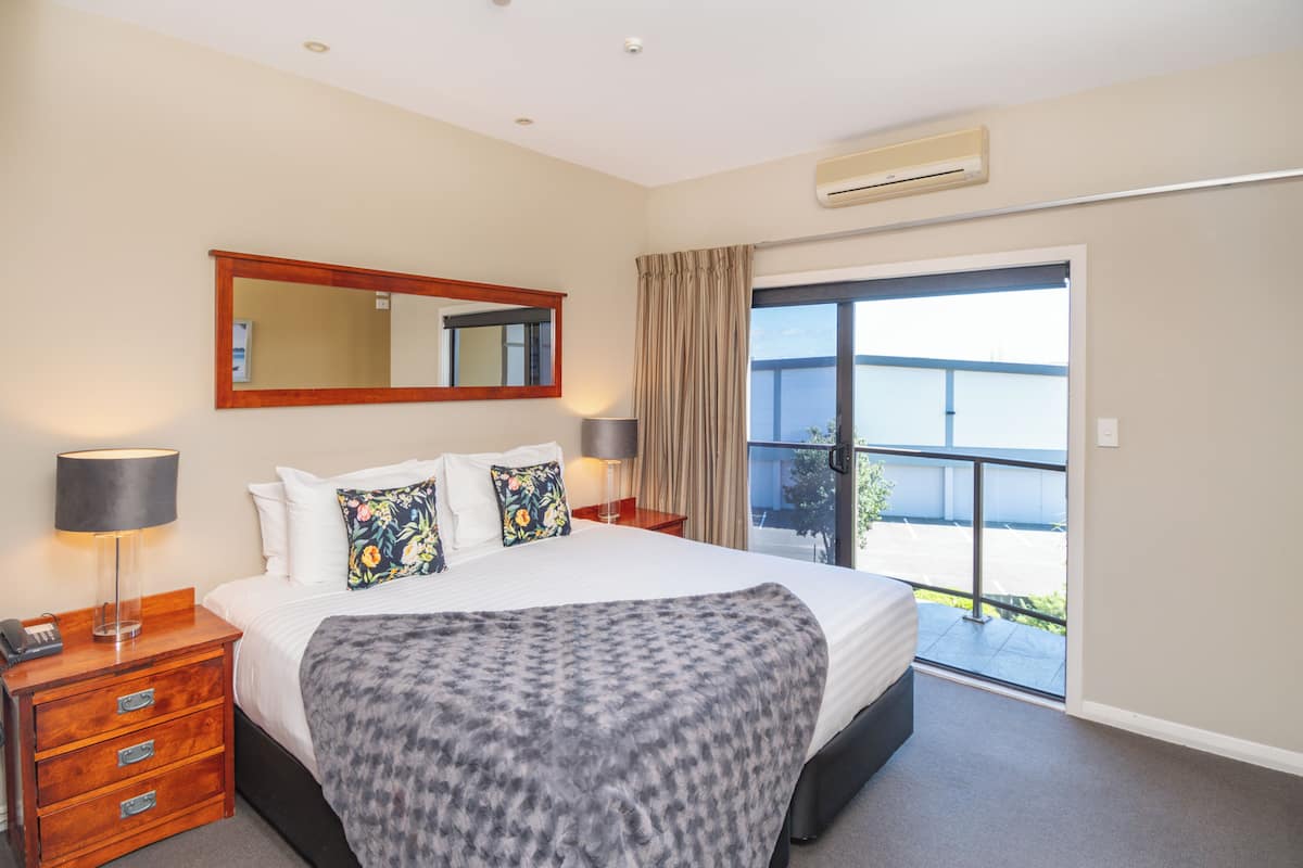 A hotel room with a large bed, a nightstand with a lamp, a mirror above the bed, and a sliding glass door leading to a balcony.