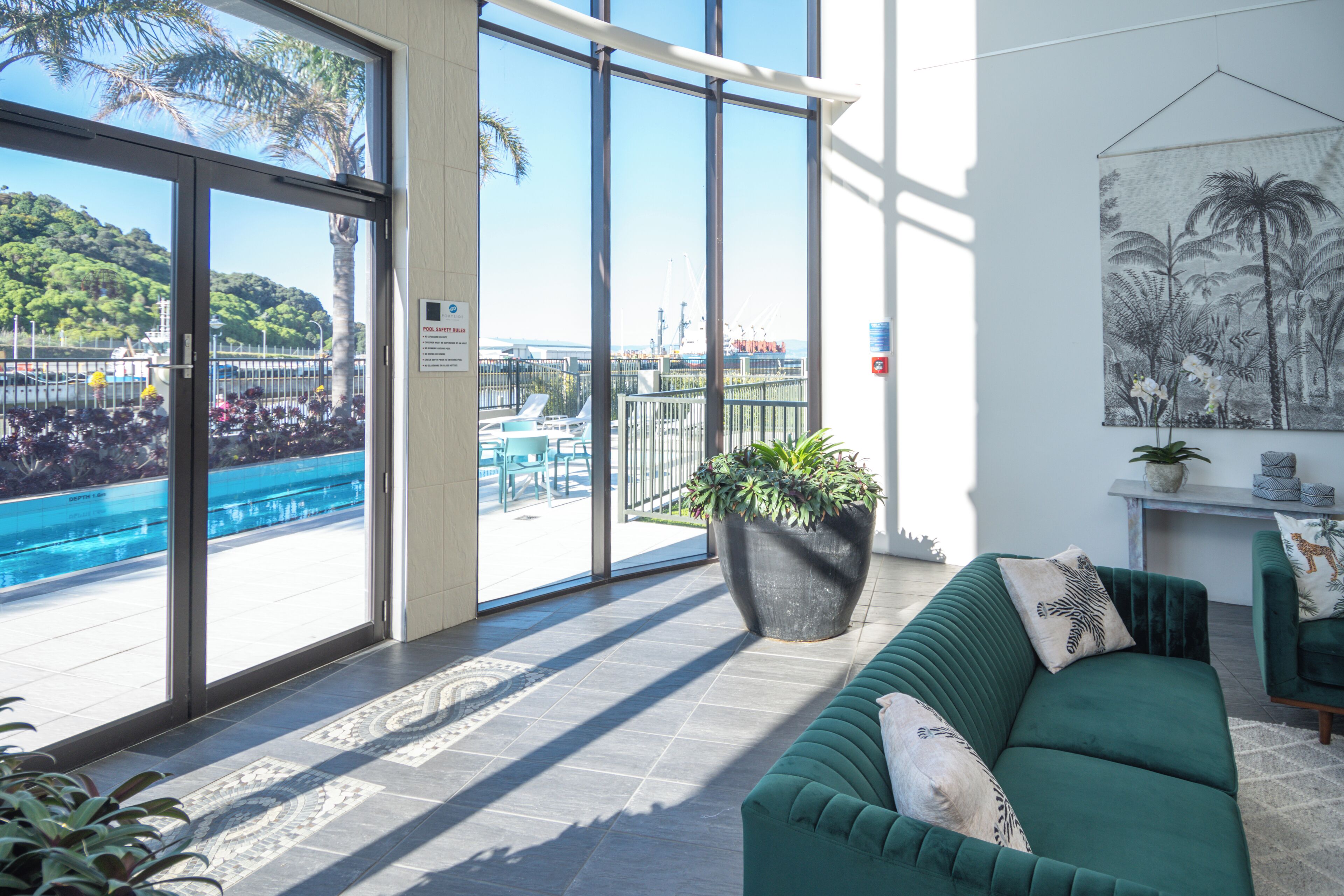 A modern hotel room with a large sliding glass door leading to a pool area, a green sofa, and a potted plant.