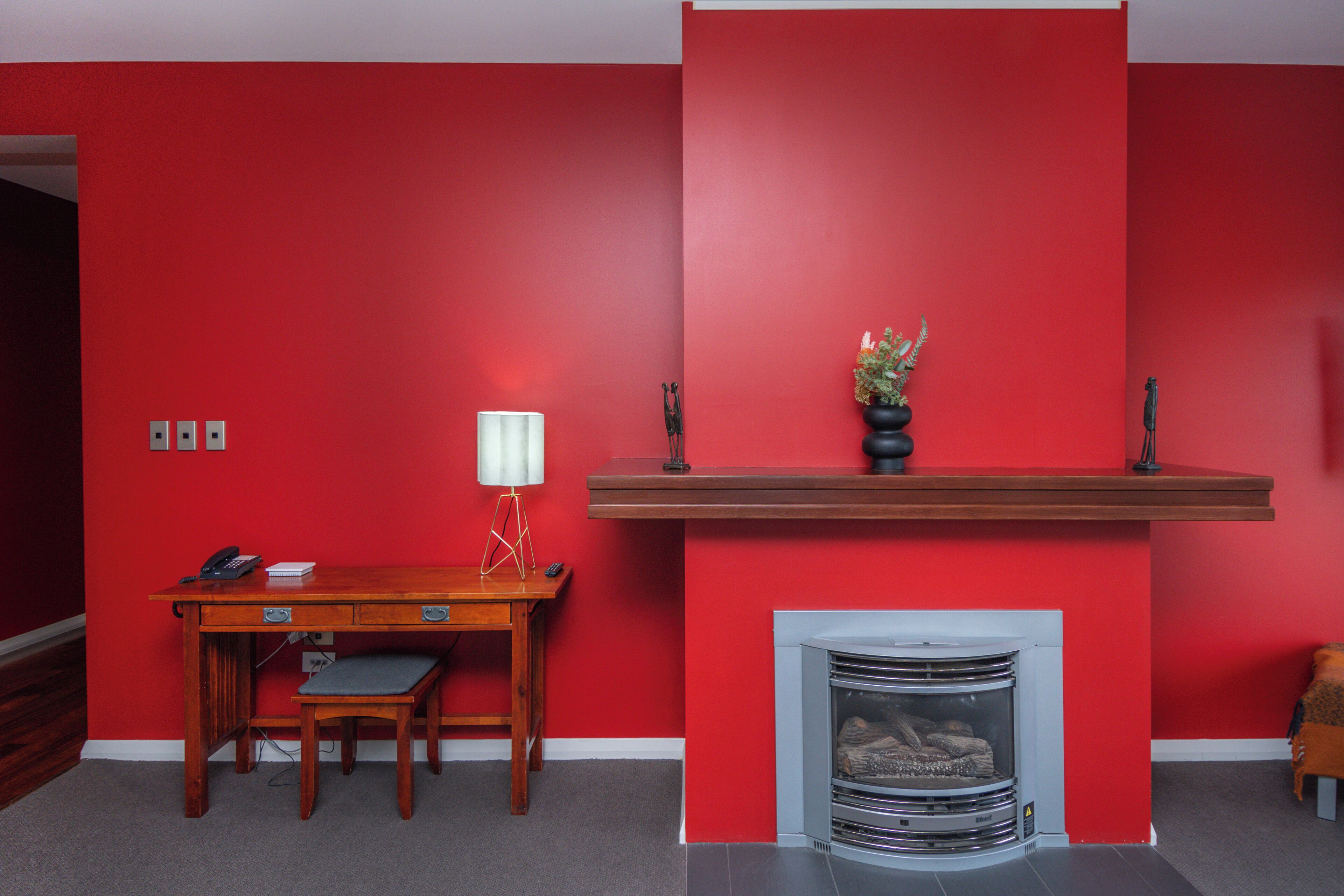 A room with a red wall, a wooden desk with a chair, a fireplace, and a lamp.
