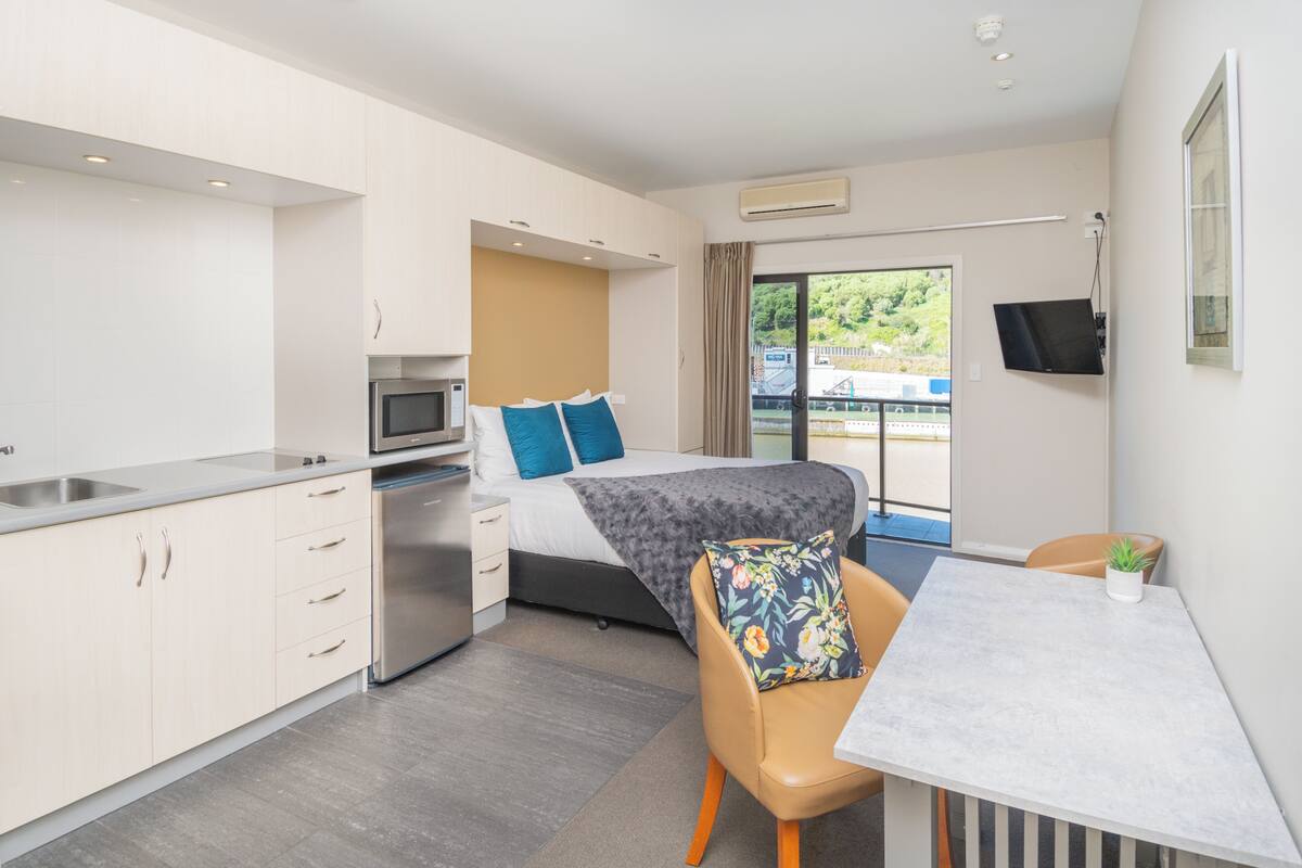 A modern hotel room with a kitchenette, a bed with blue pillows, a dining table, and a view of greenery outside.