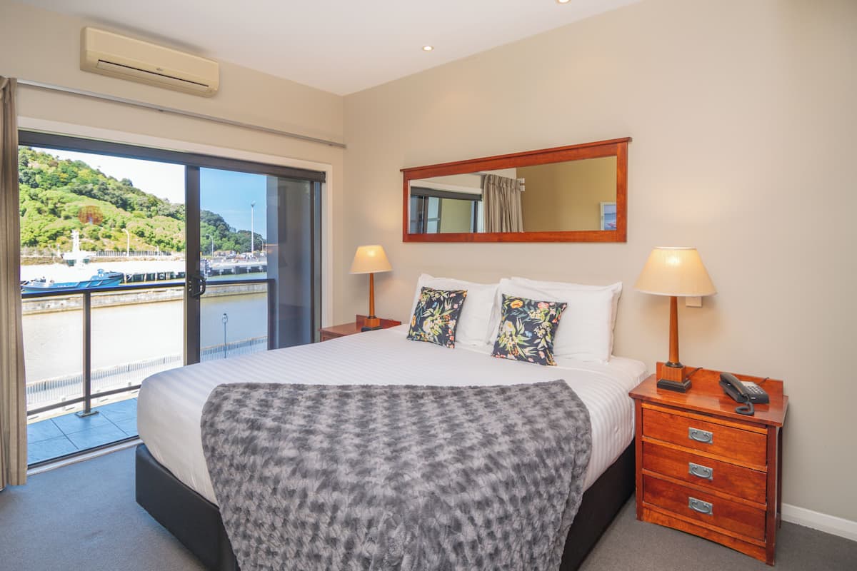 A hotel room with a large bed, a wooden nightstand with a lamp, a mirror, and a view of a body of water and greenery outside.
