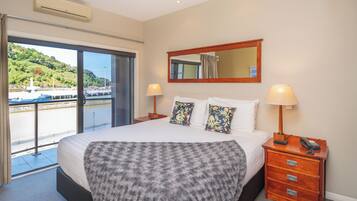Habitación de hotel con una cama grande, una mesita de noche de madera con una lámpara, un espejo y vistas al agua y vegetación.