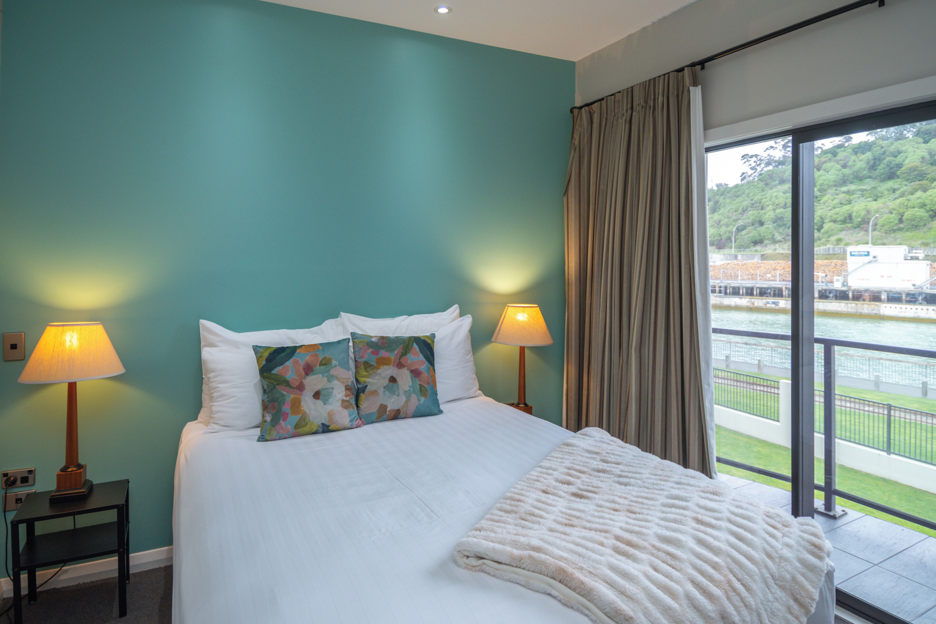 Habitación de hotel con una cama grande, dos lámparas de noche, vista a un cuerpo de agua a través de un ventanal amplio y un balcón.
