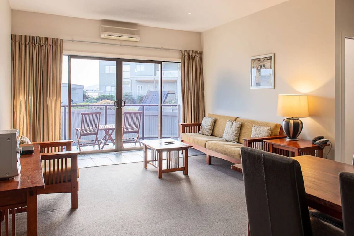 A hotel room with a sofa, chairs, a table, a lamp, and a view through a sliding door to the outside.