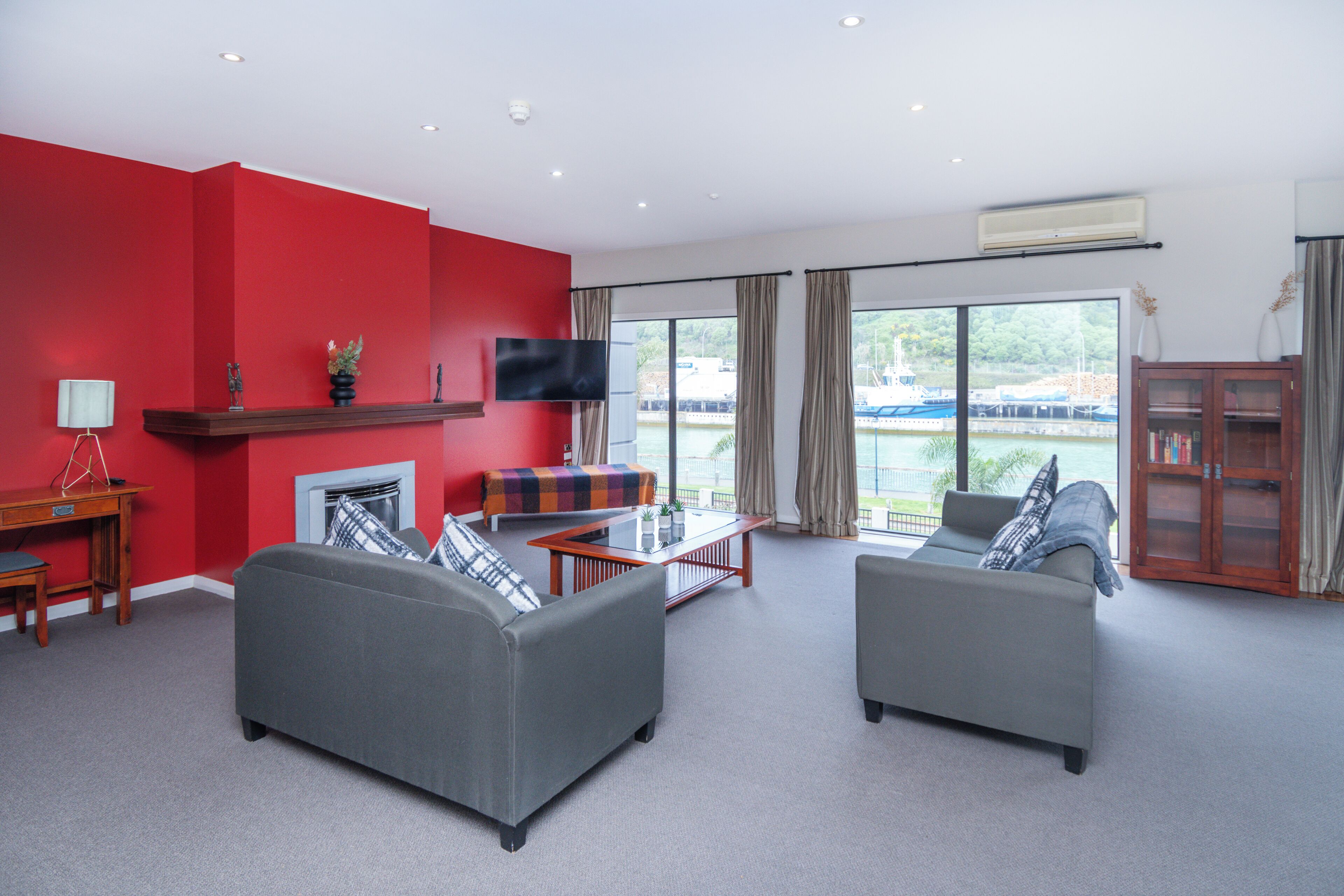 A modern living room with red walls, grey sofas, a glass coffee table, and a TV mounted on the wall.
